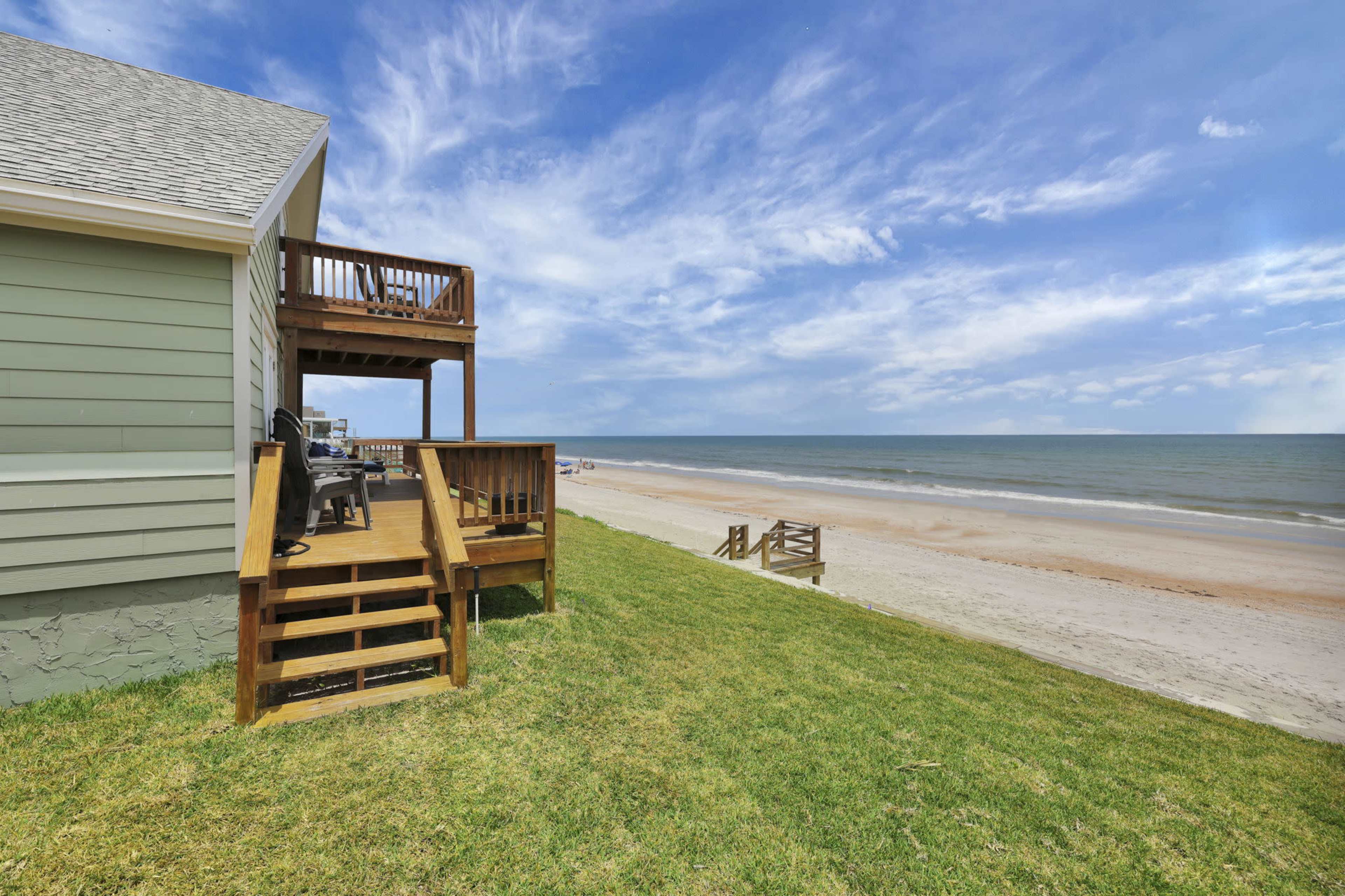 A wooden deck attached to a beach house overlooks a sandy beach and the ocean under a blue sky.