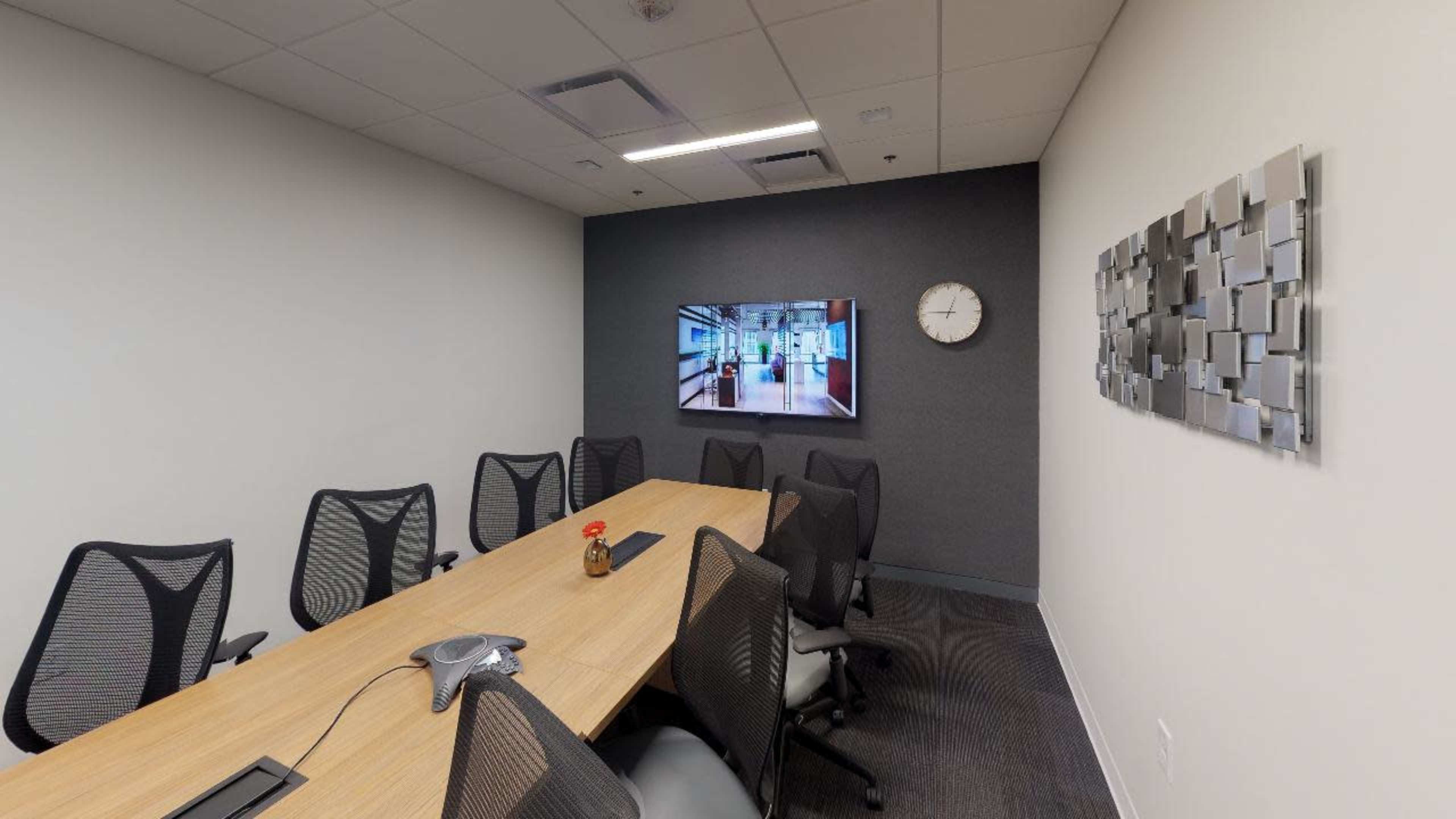 An empty conference room features a long wooden table surrounded by ergonomic chairs, a large wall-mounted screen, and a clock.