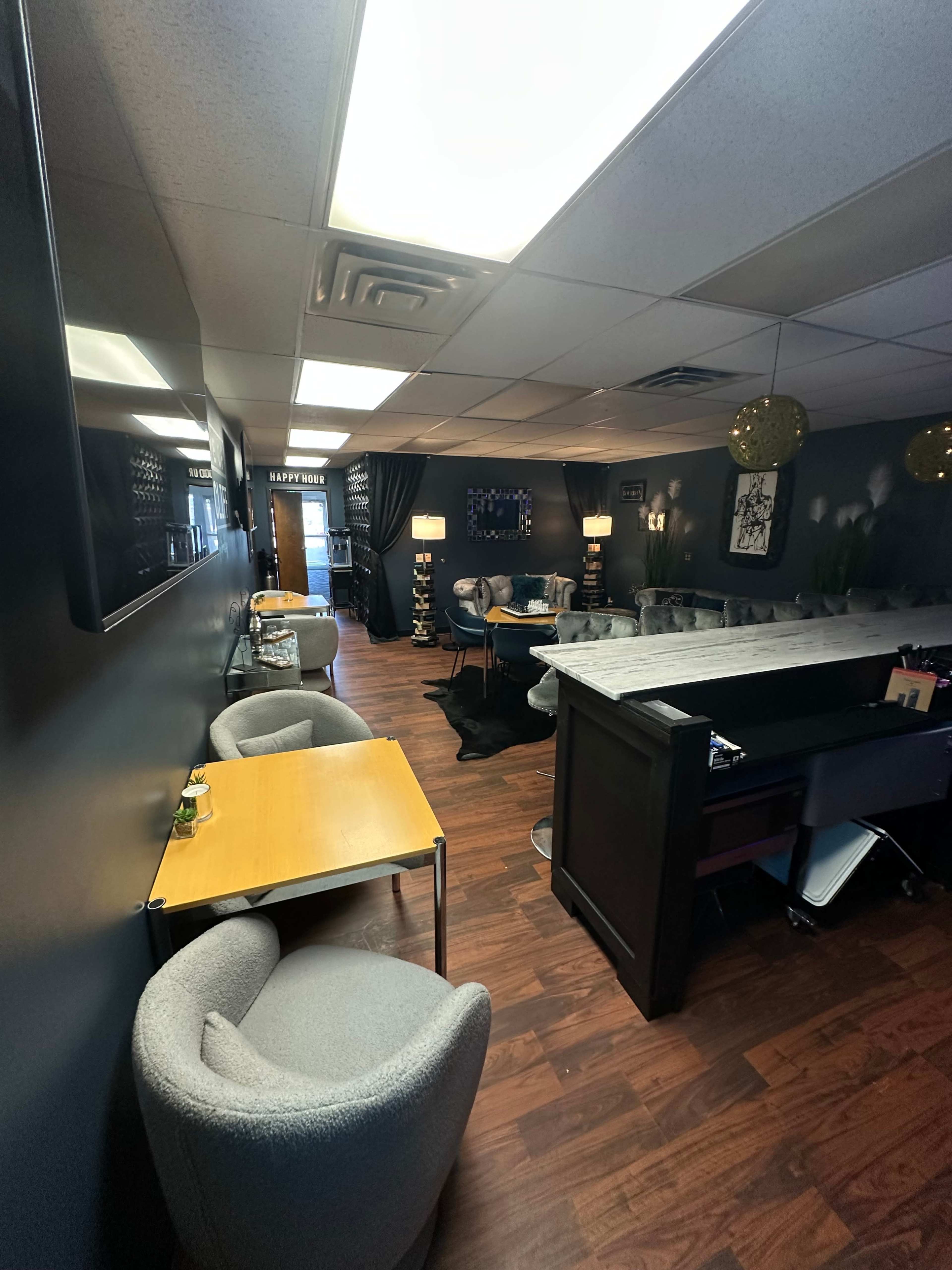 The image shows a modern interior of a salon or lounge with gray upholstered chairs, small tables, and a bar area, all under bright ceiling lights.