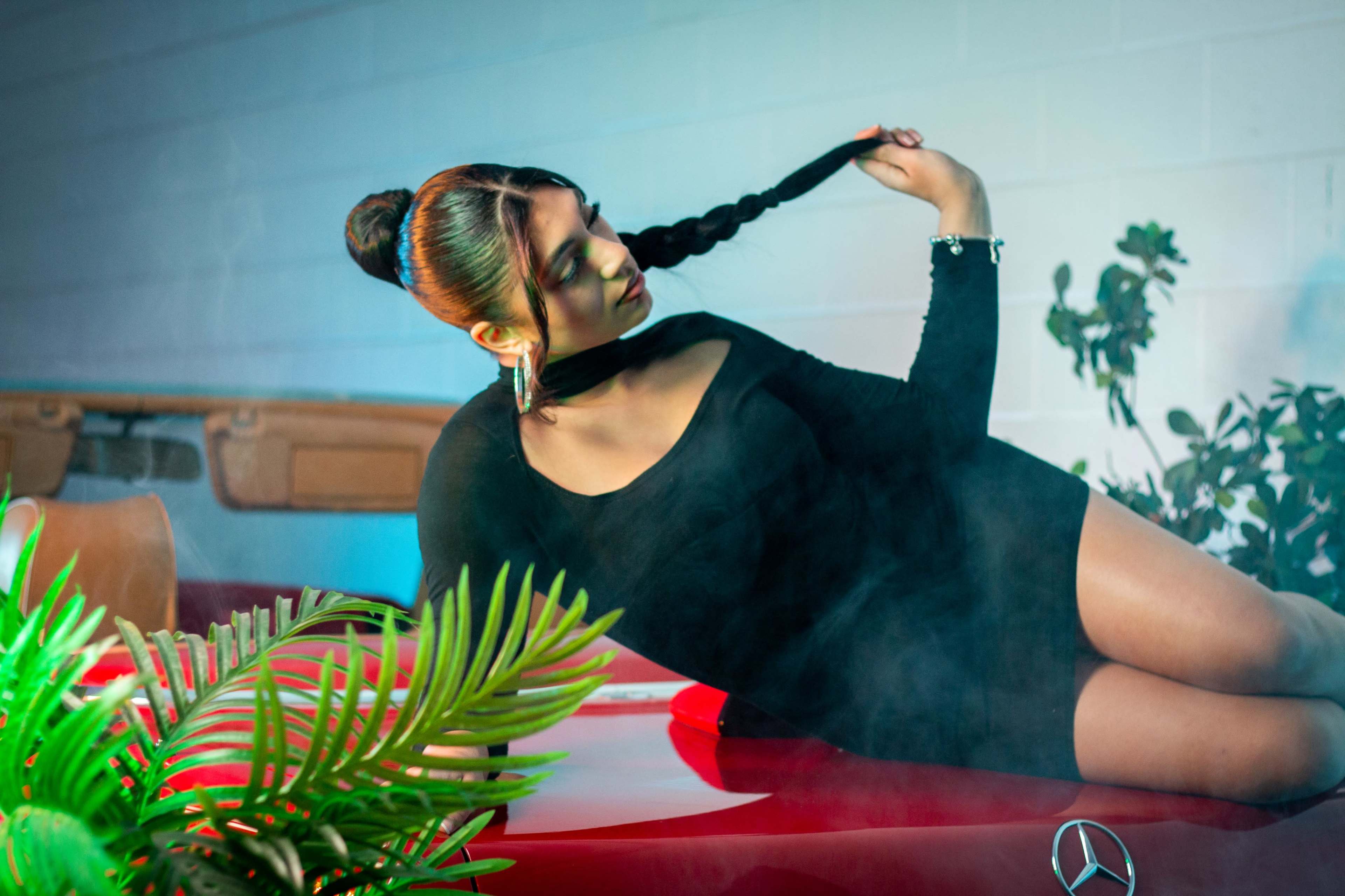 A woman in a black dress poses on the hood of a red car under a smoky atmosphere, with green plants in the foreground.