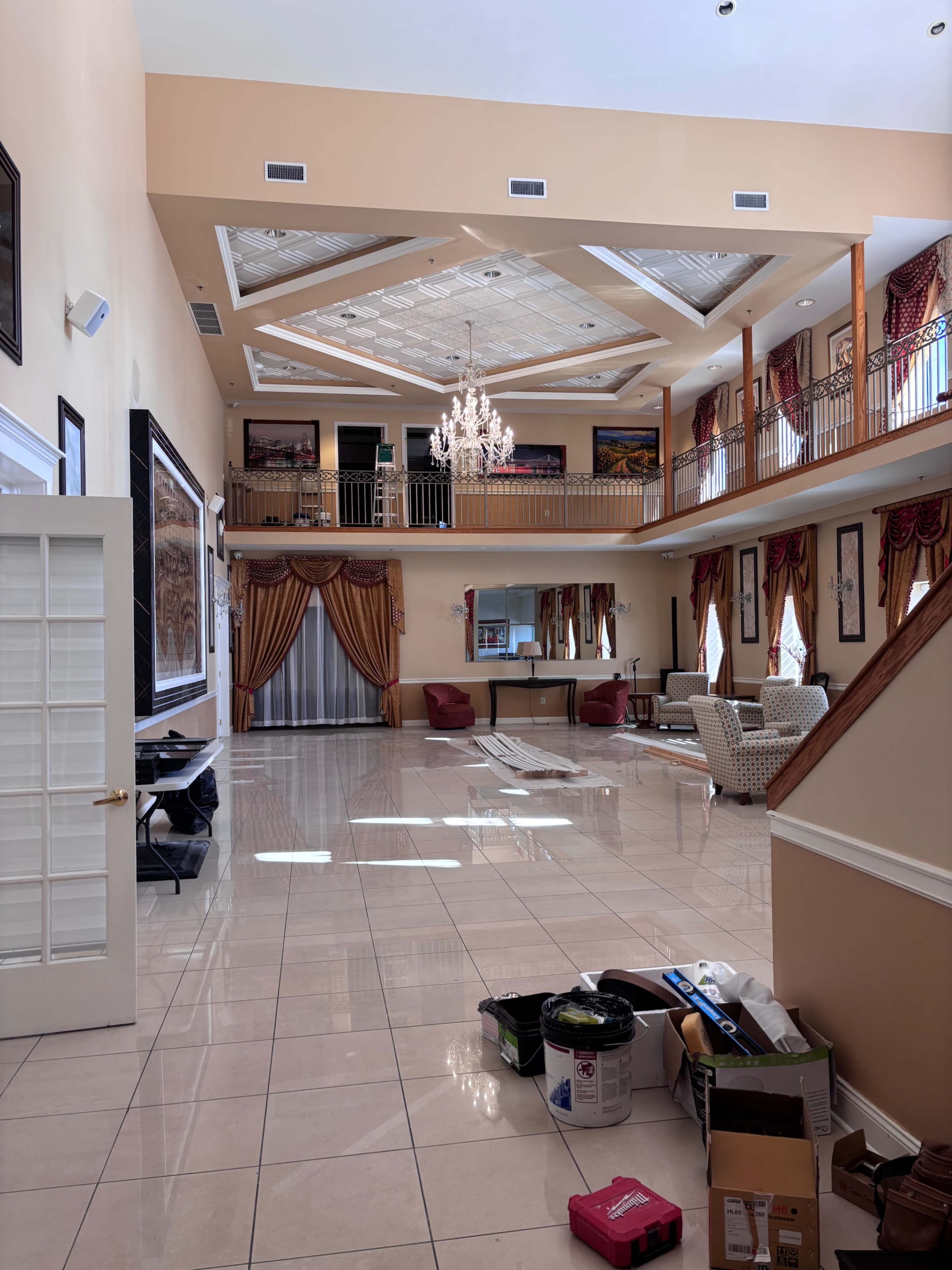 The image shows a large, open foyer with high ceilings, a chandelier, and two levels featuring a staircase and various pieces of furniture.