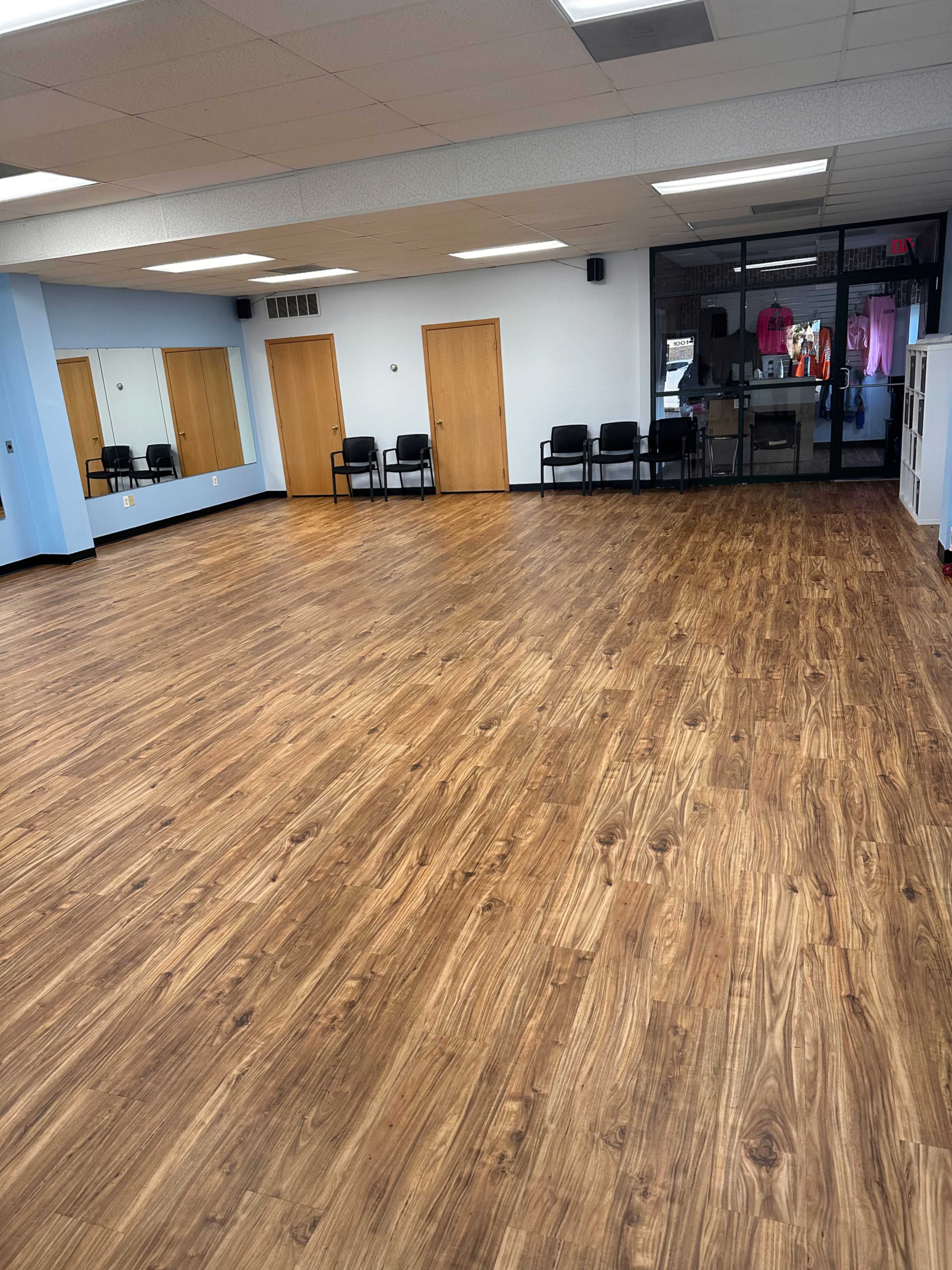 A large, empty room with wooden flooring, mirrors on one wall, and several black chairs arranged on one side.