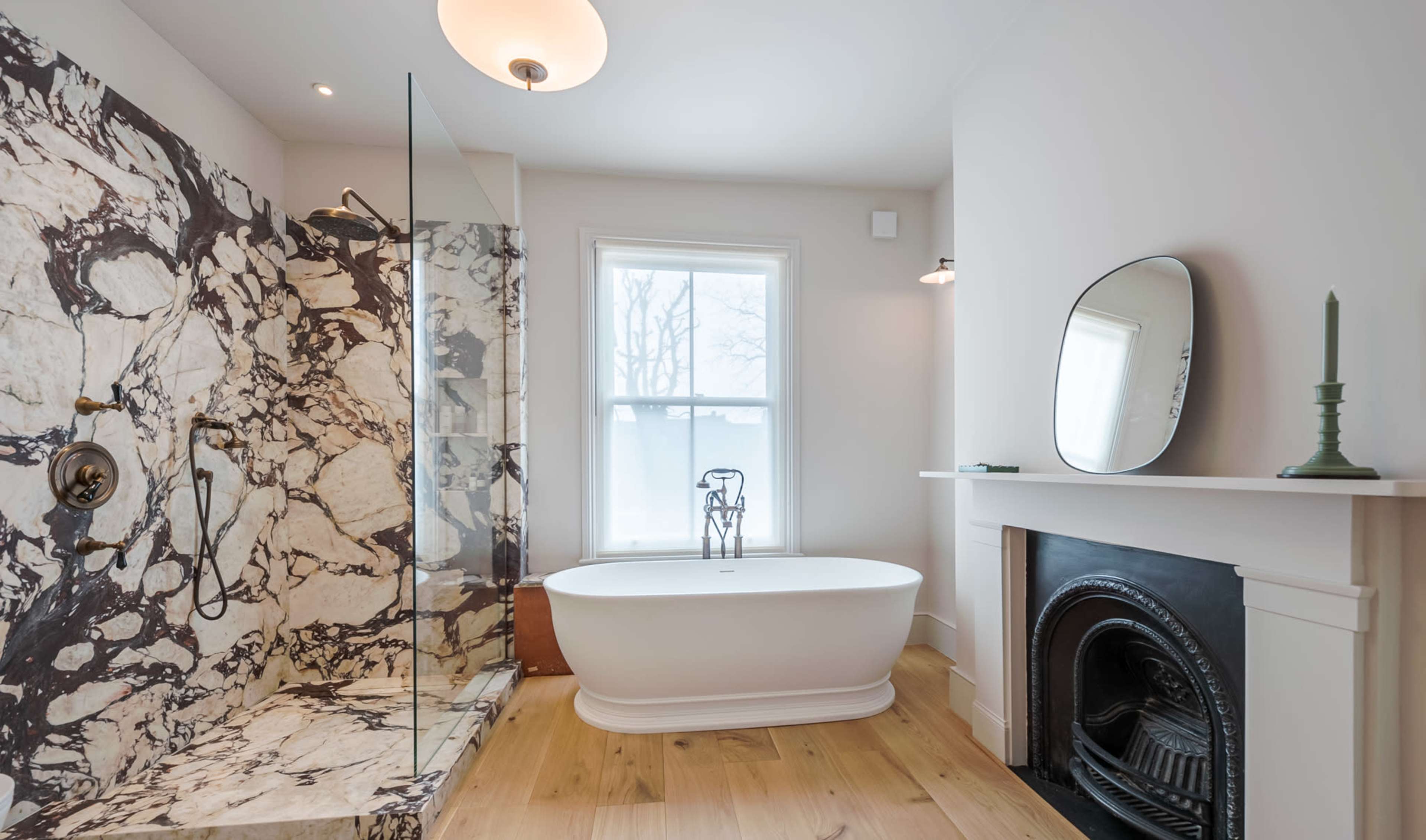 The image shows a modern bathroom featuring a freestanding tub, a marble shower enclosure, and a decorative fireplace.