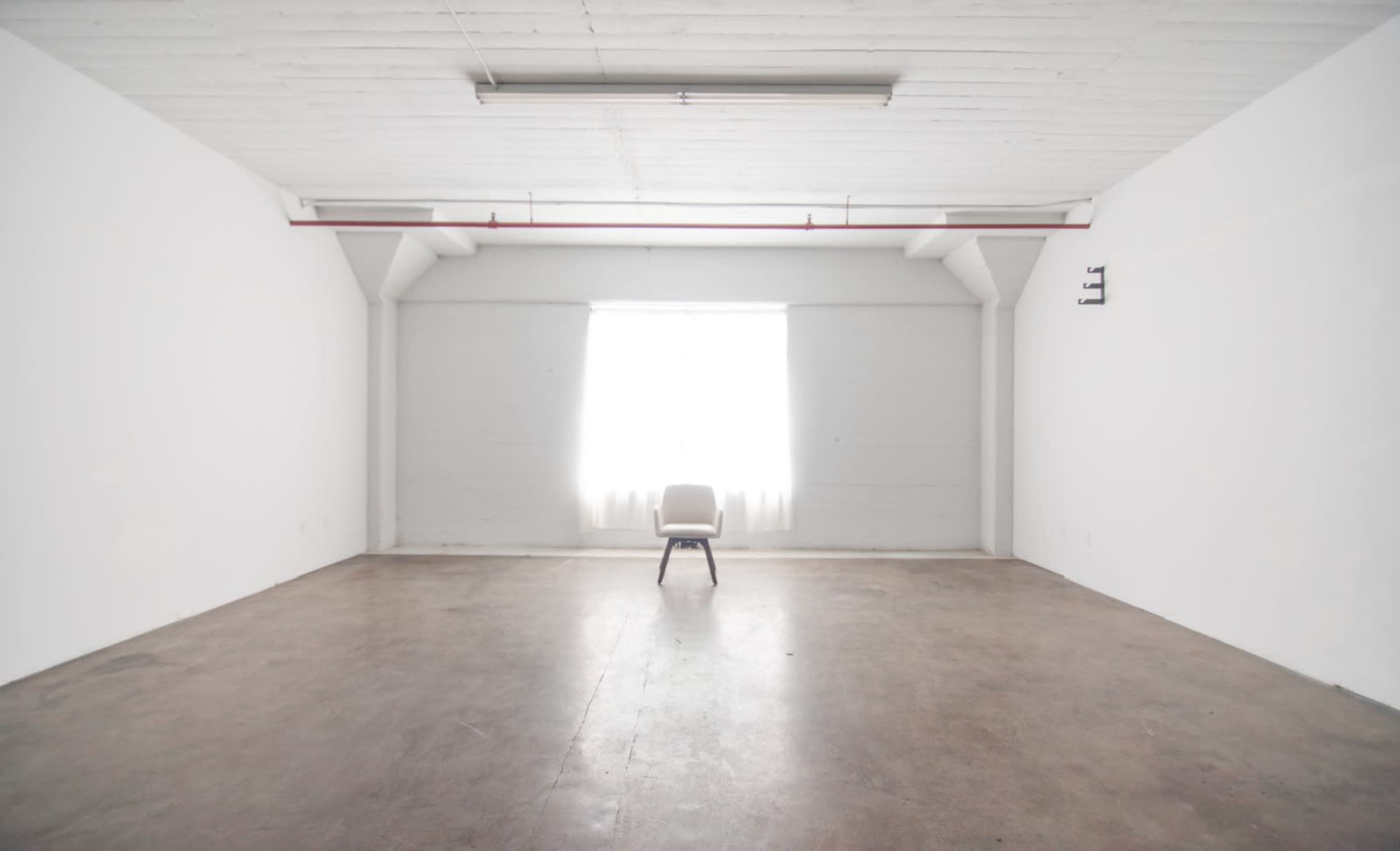 A single chair is positioned in the center of a vast, empty room with white walls and a bright window.