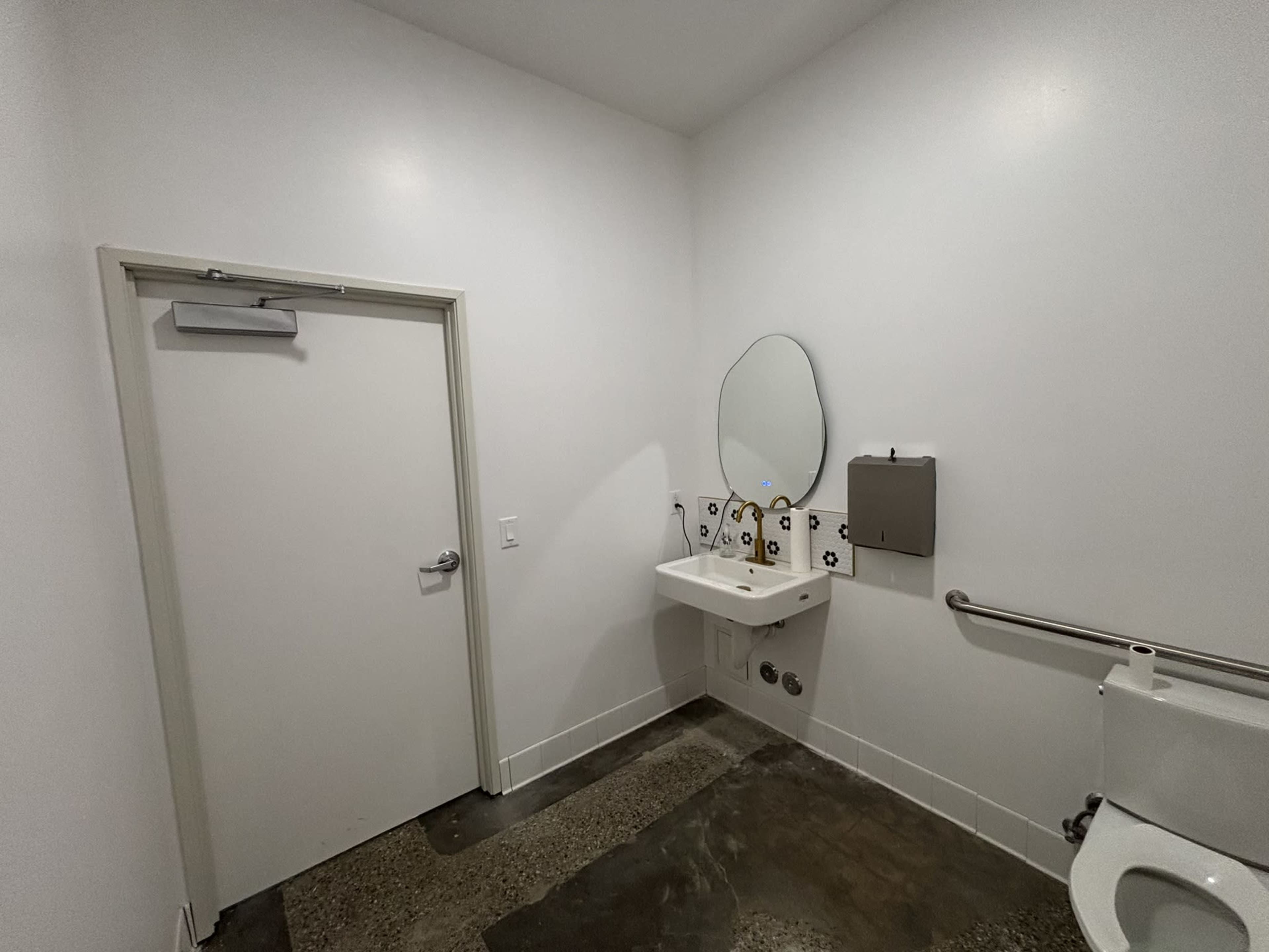 The image shows a clean, minimalist restroom with a single toilet, a small sink with a mirror above it, and a door on one side.