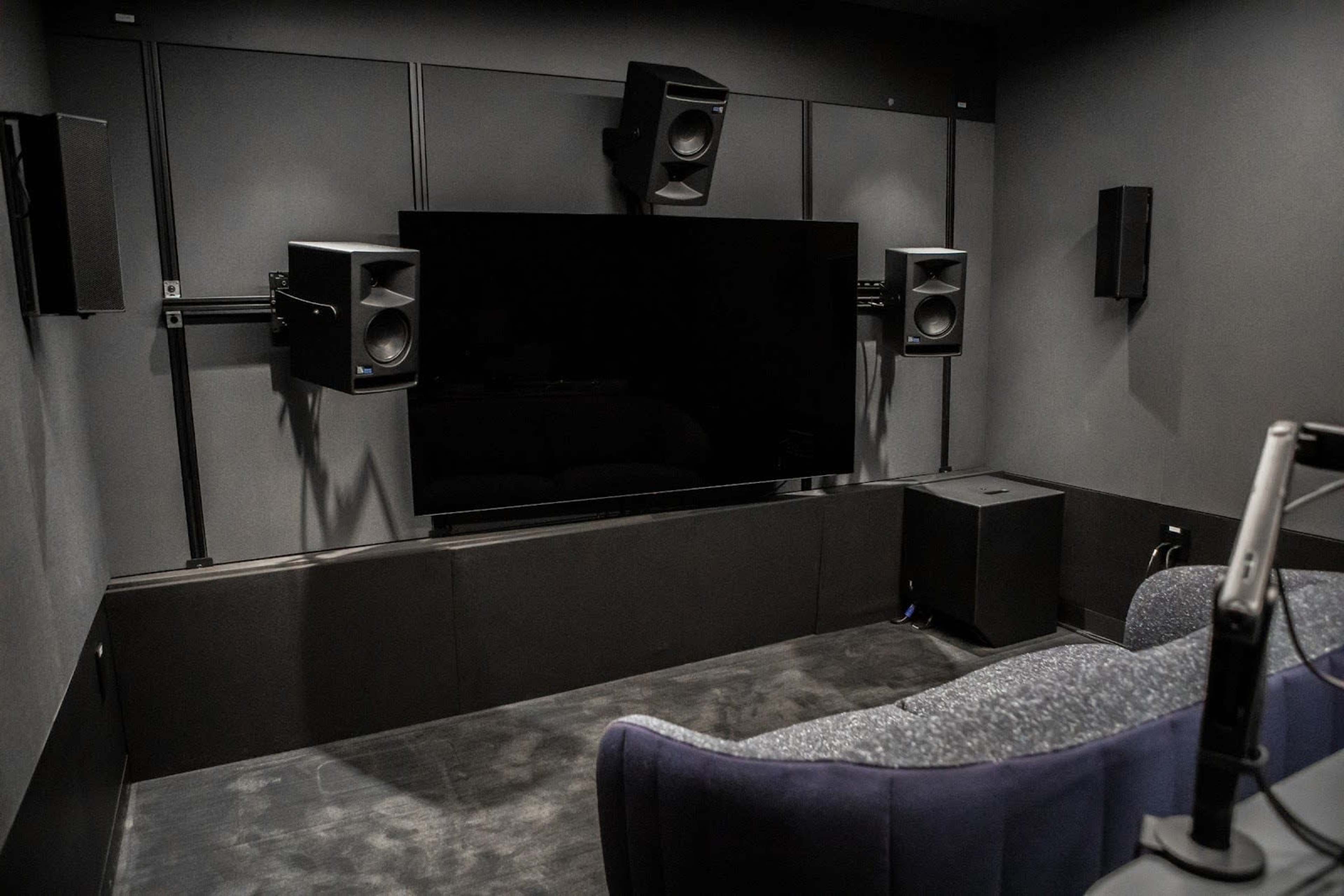 The image shows a home theater room with a large flat-screen TV mounted on the wall, surrounded by multiple speakers and a plush seating area.