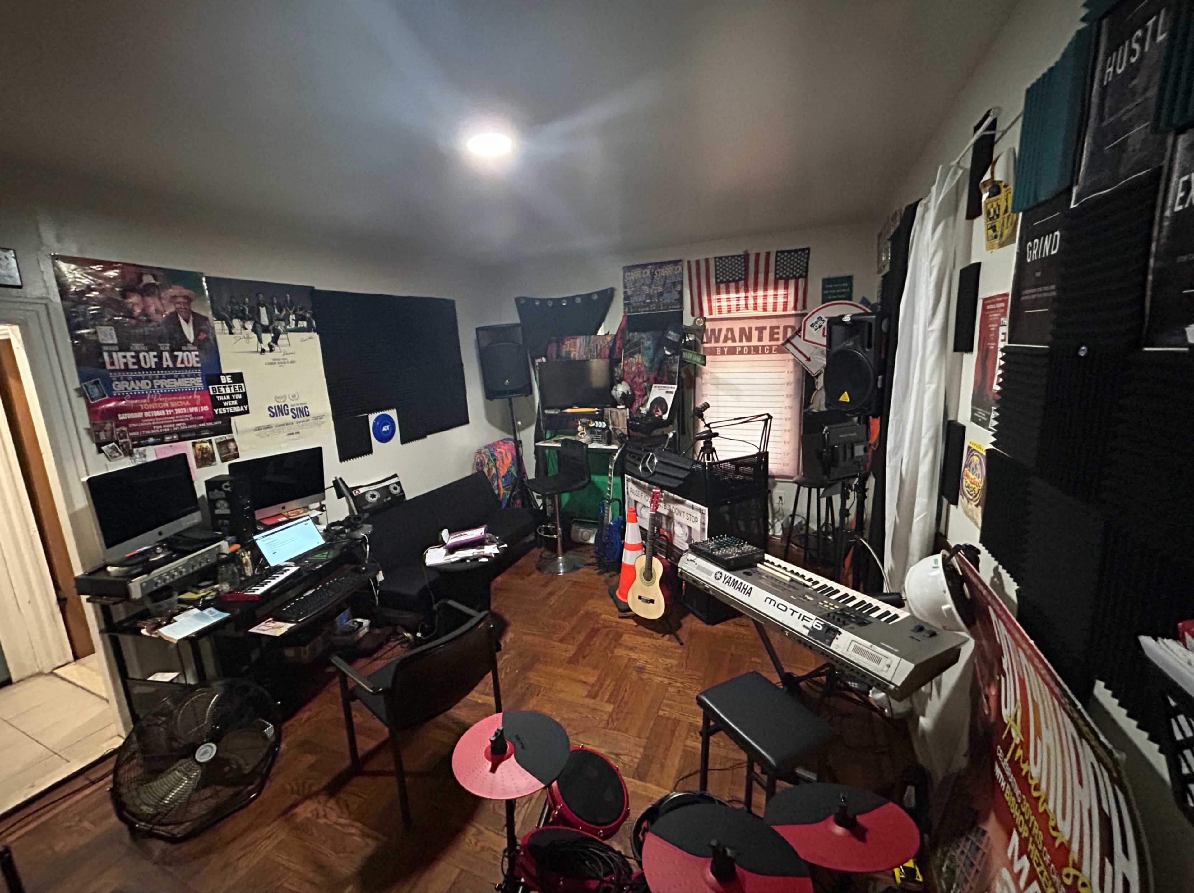 The image shows a small music studio with various instruments, sound equipment, and posters on the walls.