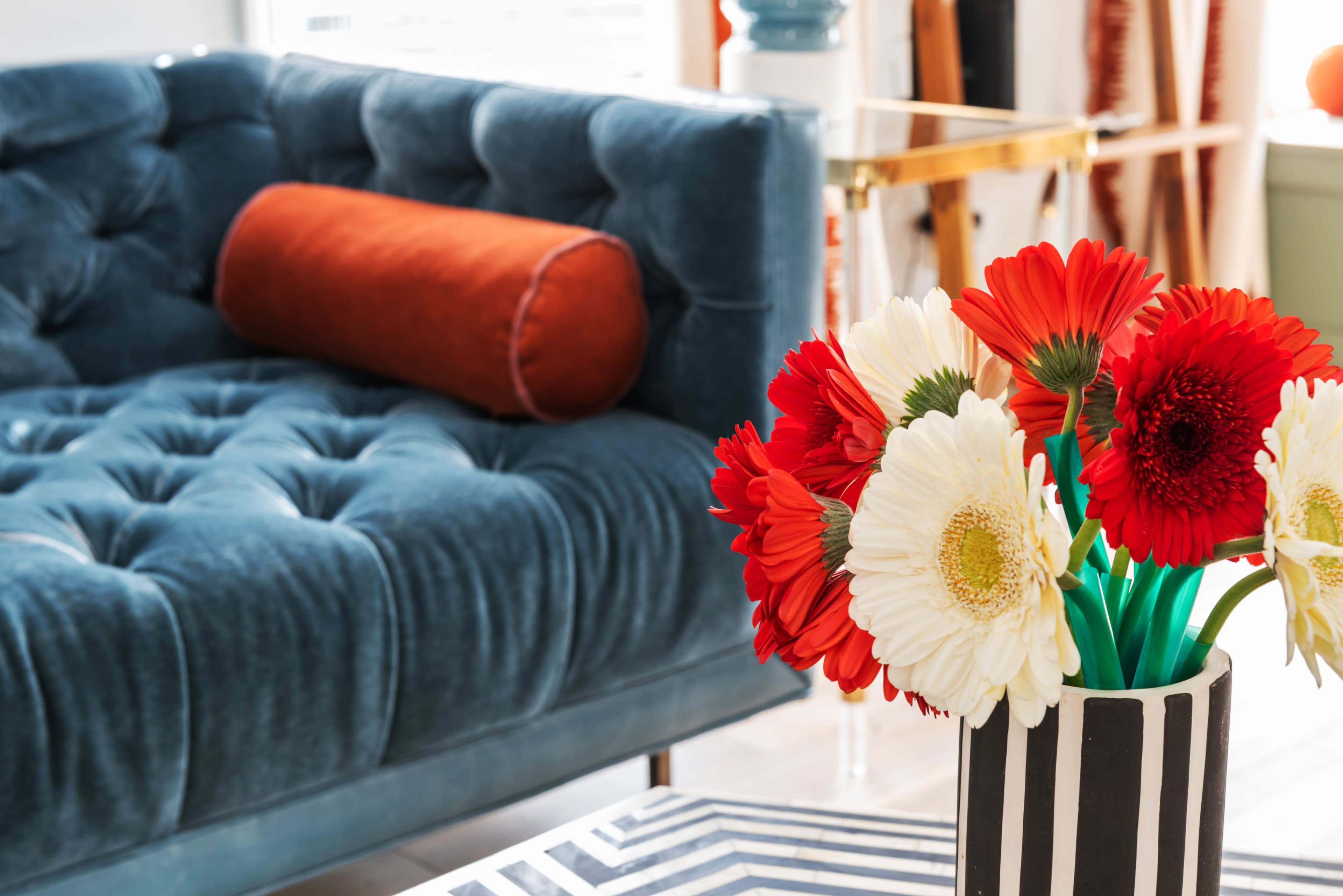 A blue tufted sofa is paired with a striped table featuring a vase of red and white flowers.