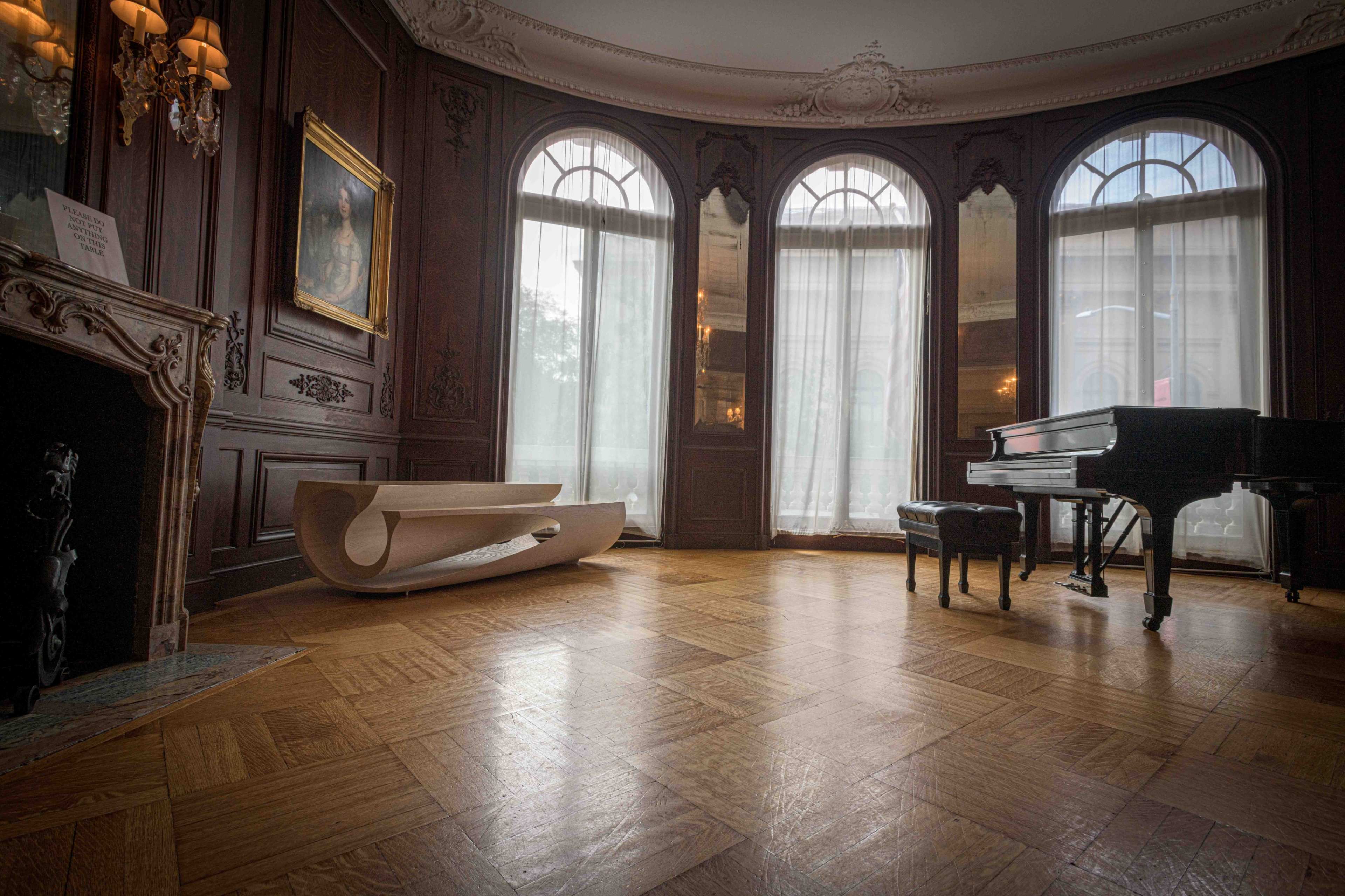 A room with dark wooden walls, large arched windows, a grand piano, and a curved wooden bench on a patterned wooden floor.