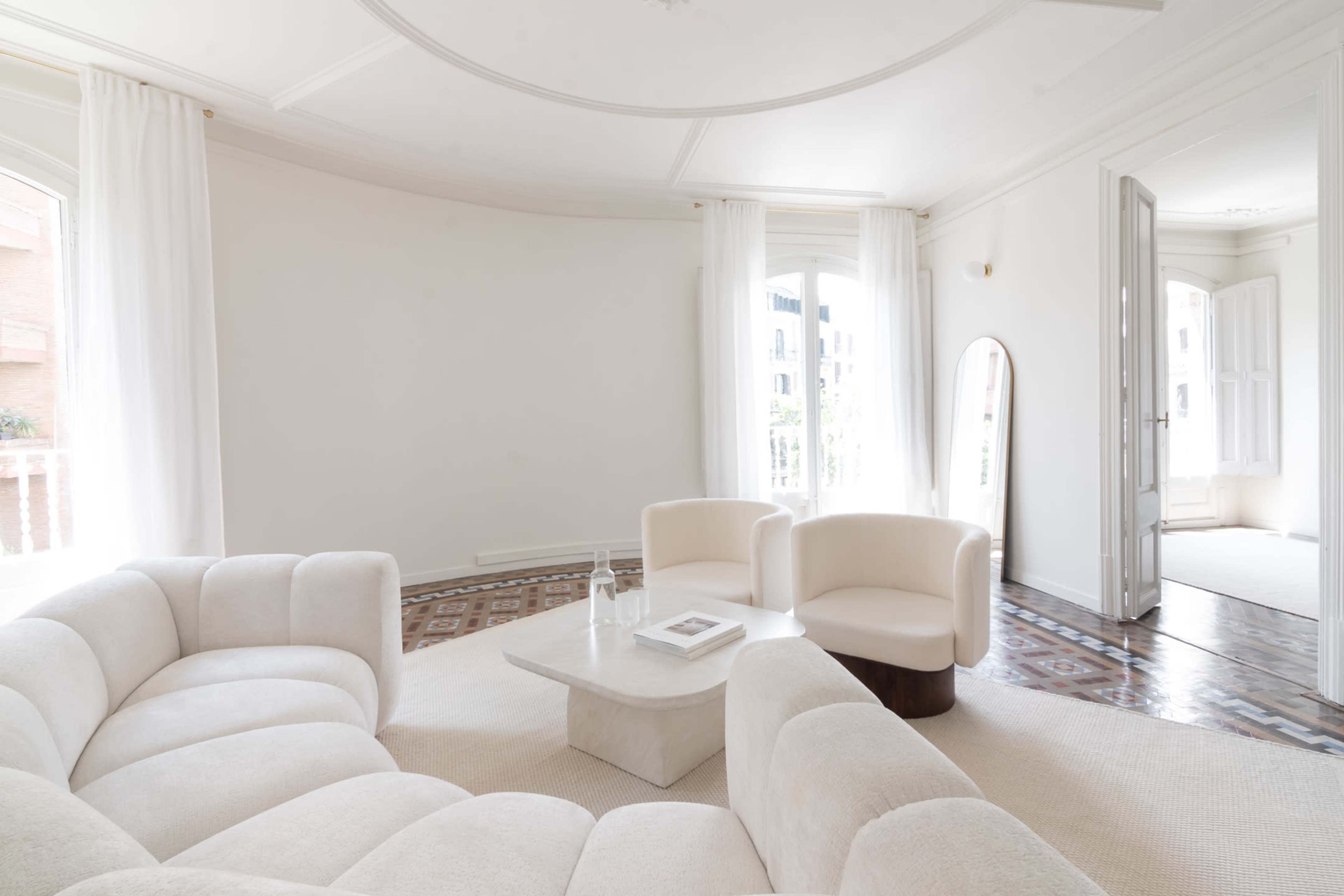 The image shows a bright, minimalist living room with a curved sofa, a coffee table, and a large mirror, featuring wooden floors and light curtains.