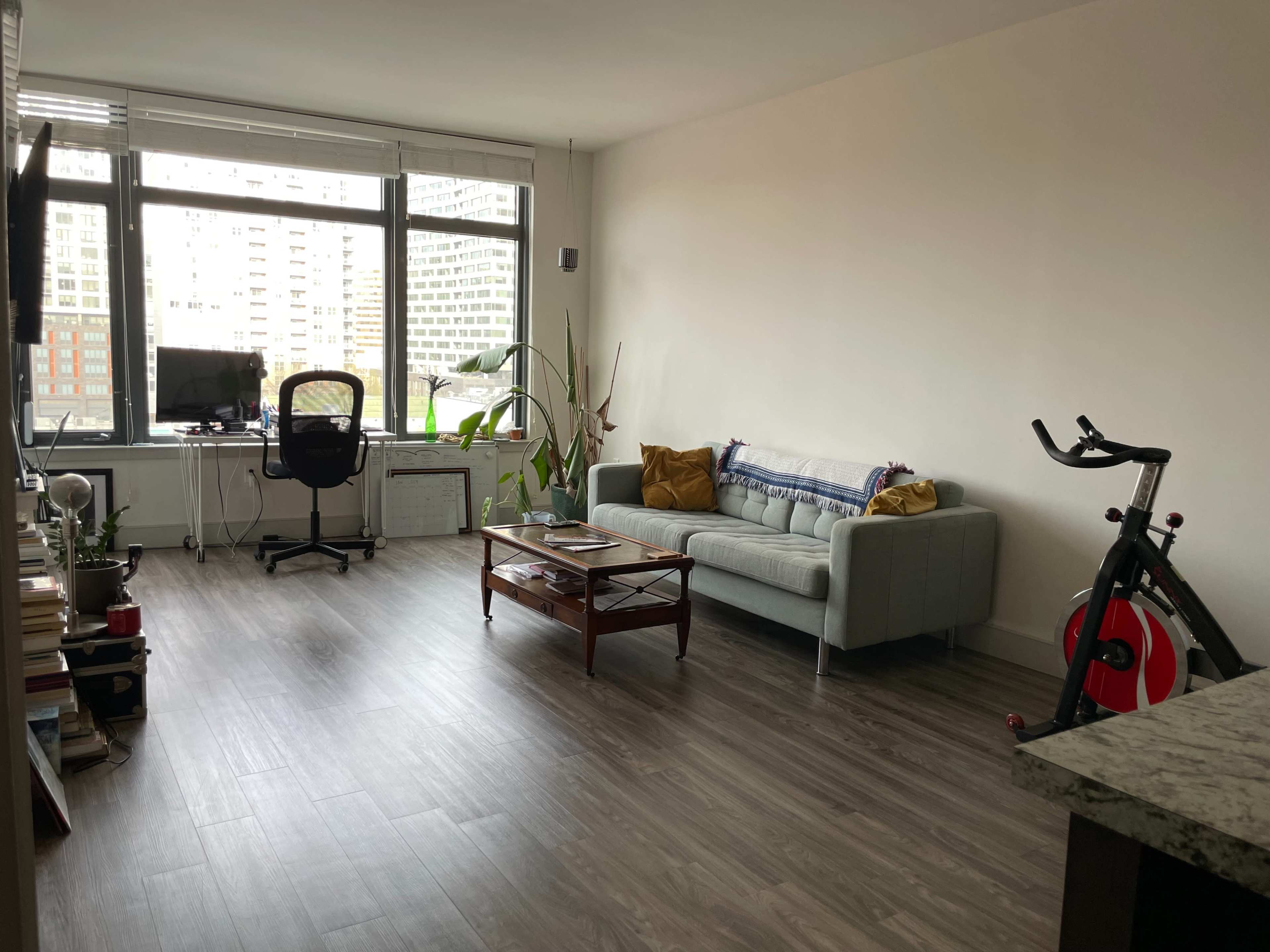 A spacious living area with a light gray couch, a wooden coffee table, a black office chair, a bicycle, and large windows with city views.