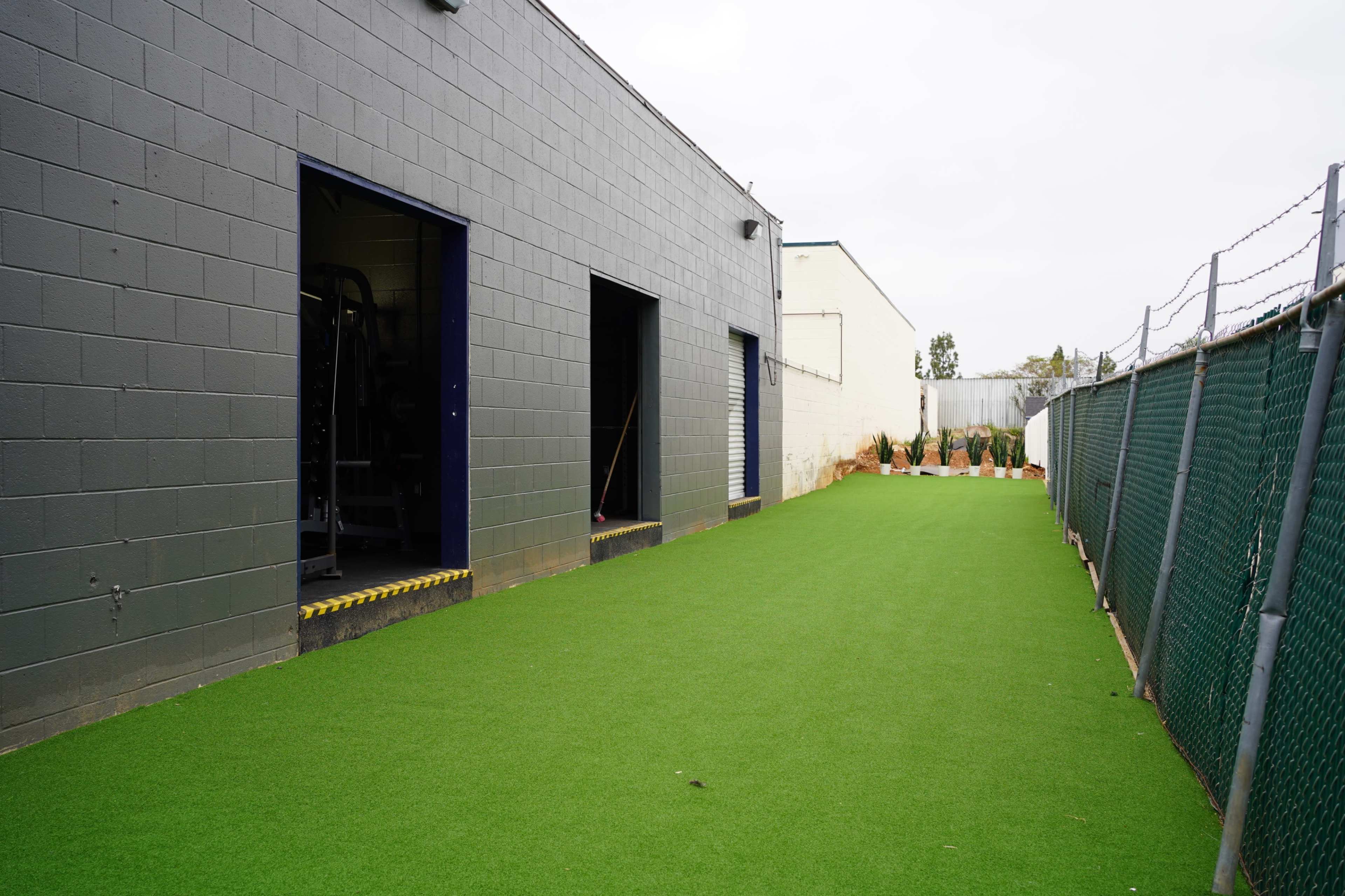 A narrow outdoor space with artificial turf beside a gray building and an adjoining fenced area.