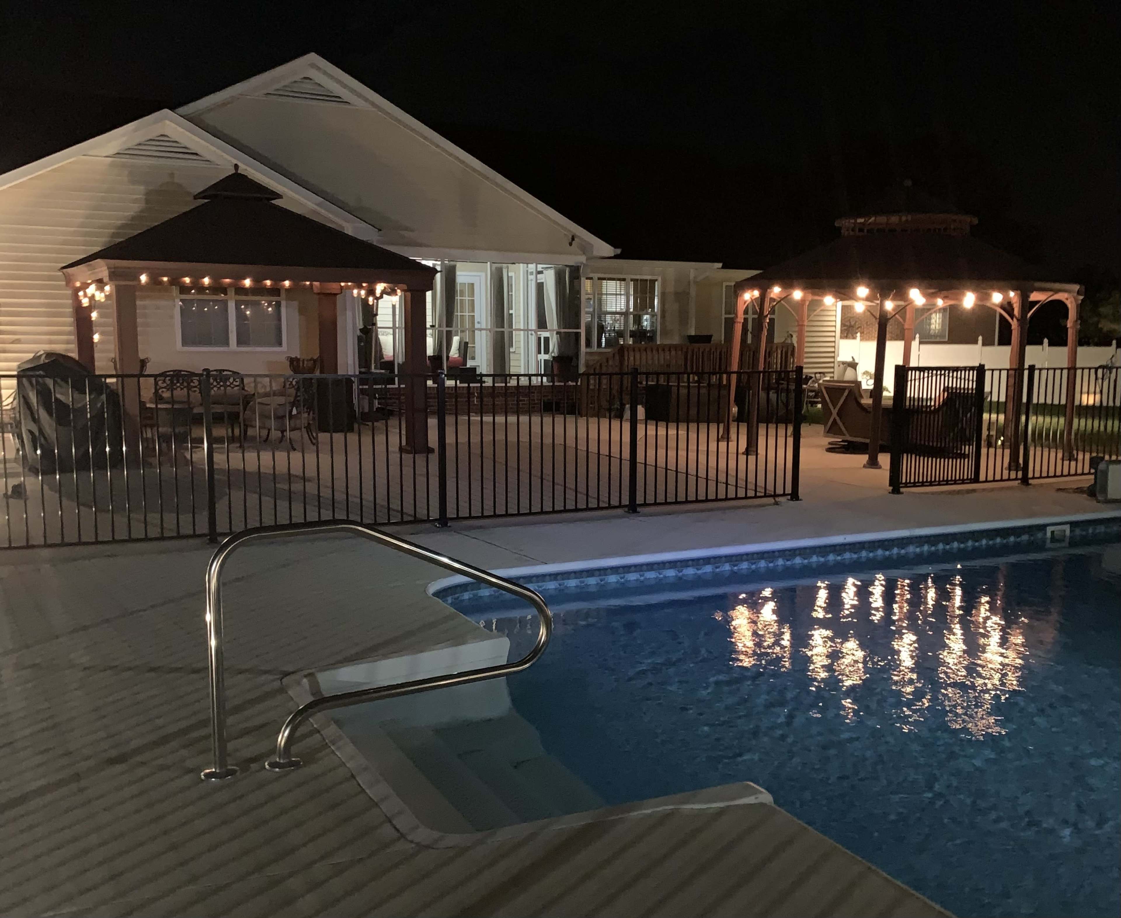 The image shows a well-lit backyard at night, featuring a swimming pool with a ladder, a fenced patio area with two gazebos, and a house in the background.