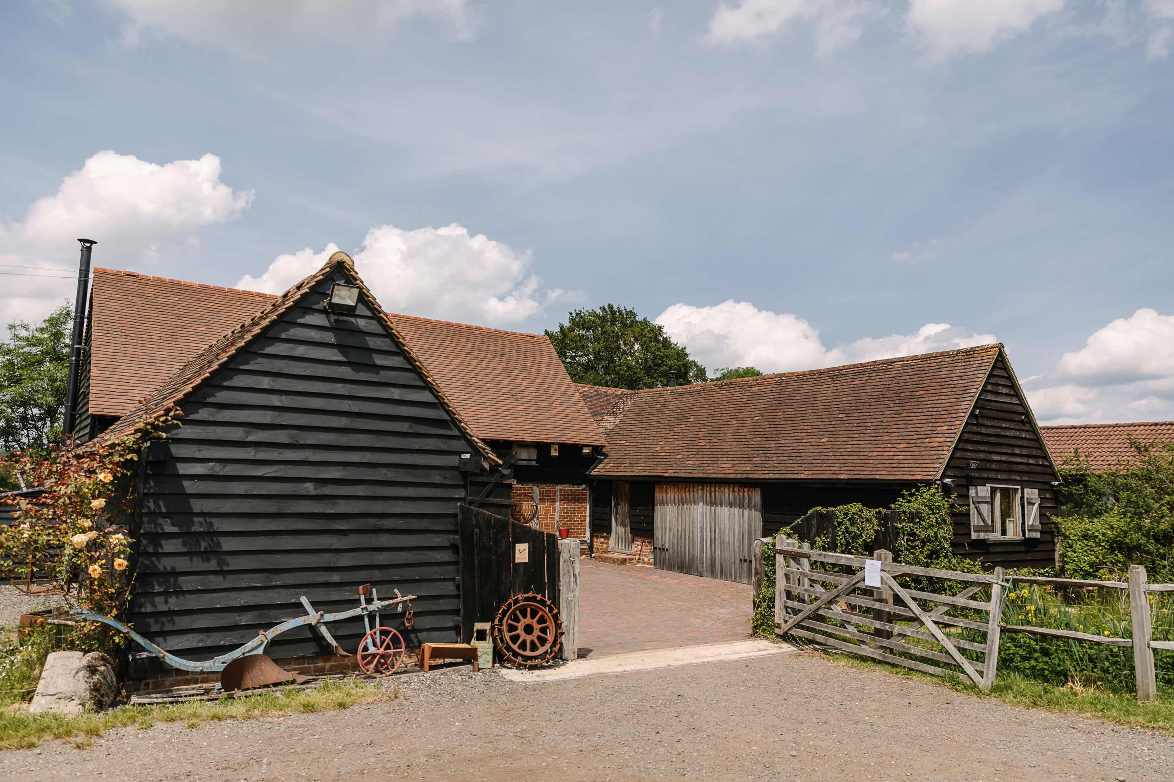 A black wooden barn with a red-tiled roof is situated beside a gravel pathway, accompanied by a vintage cart and wooden wheel.