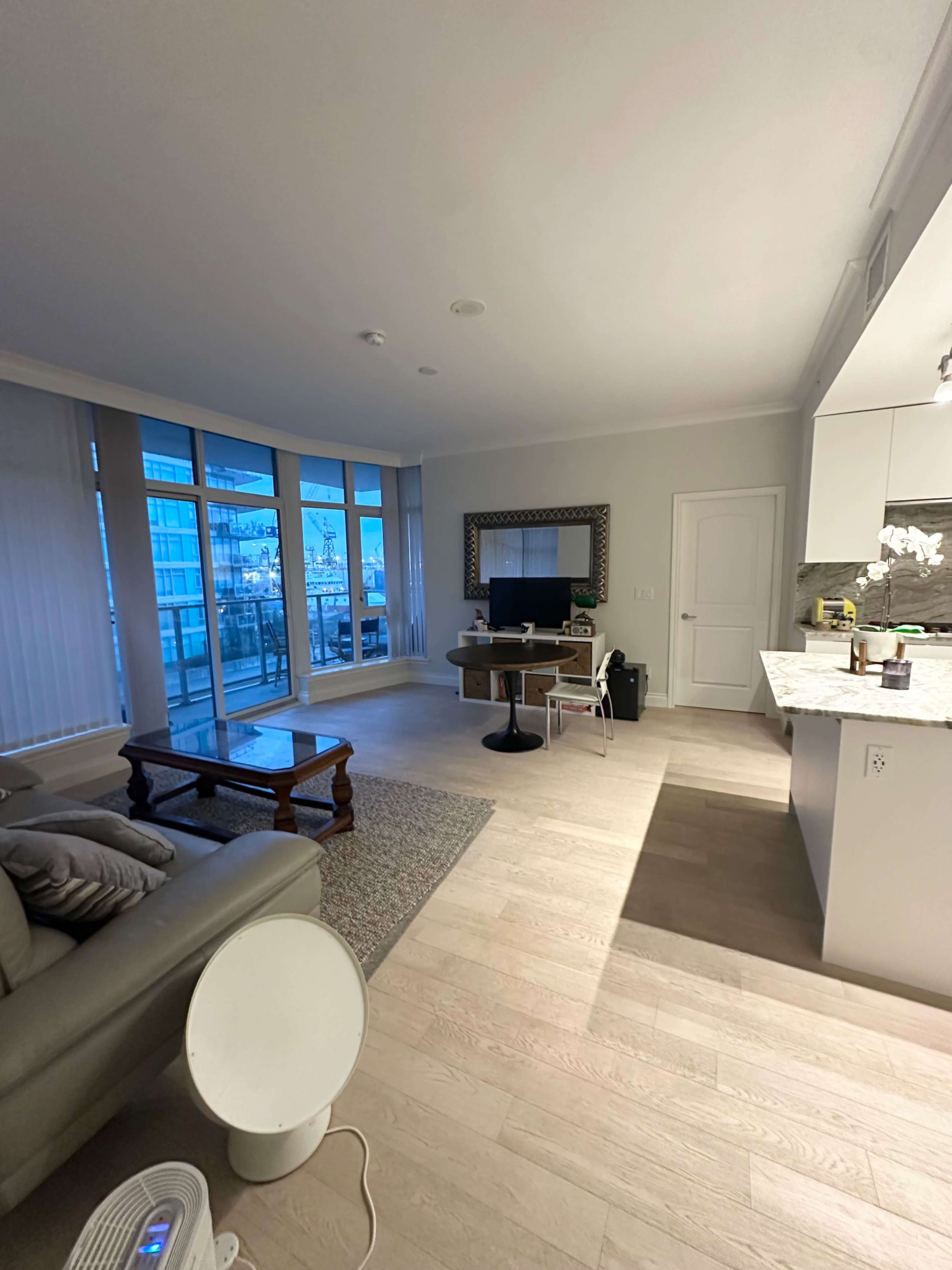 The image shows a modern living room with a sofa, dining table, television, and large windows allowing natural light to enter.