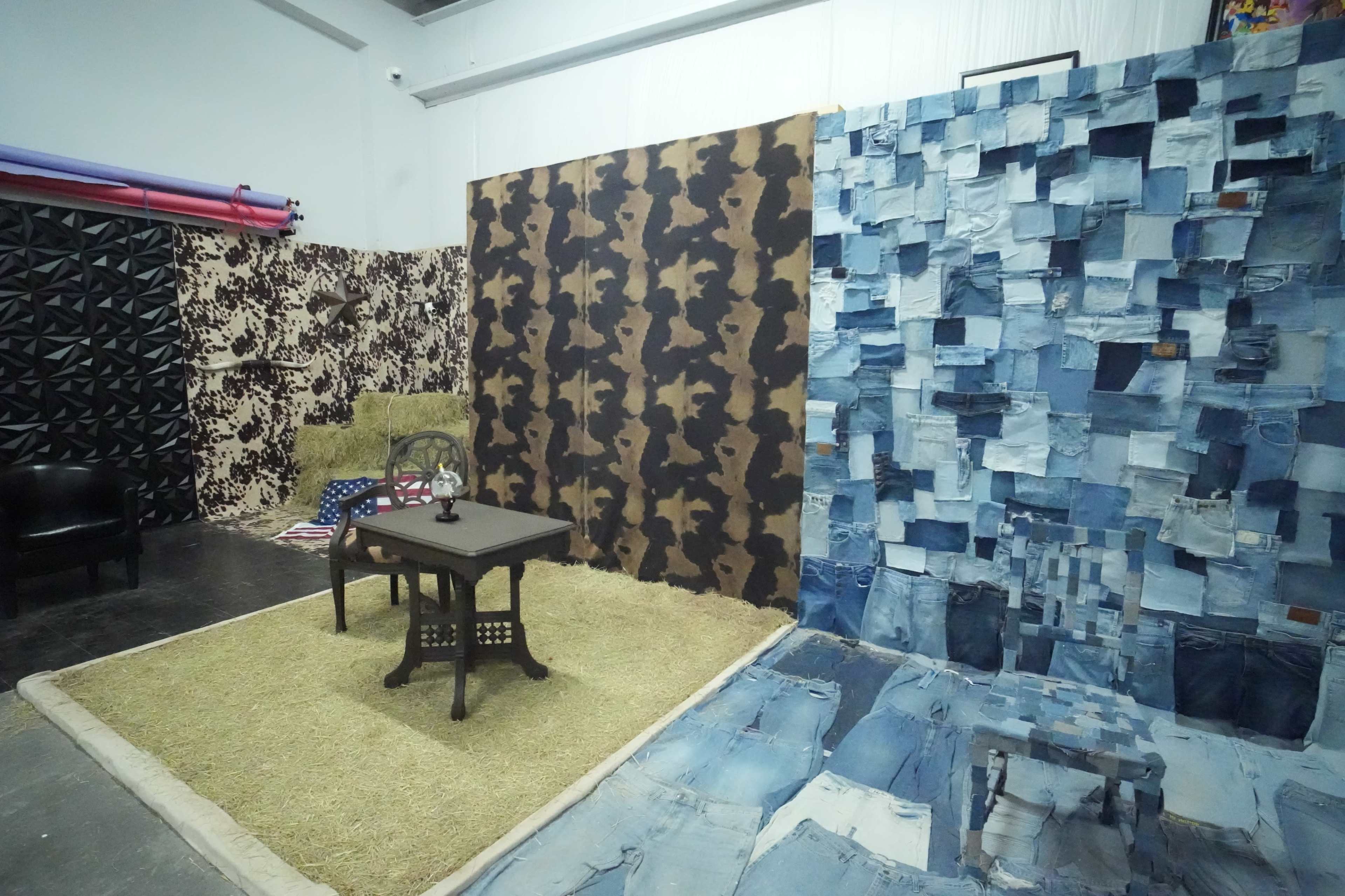 The image shows a room with four textured walls, featuring a mix of denim, camouflage patterns, and fabric, with a small table and chair set on a green rug in the center.