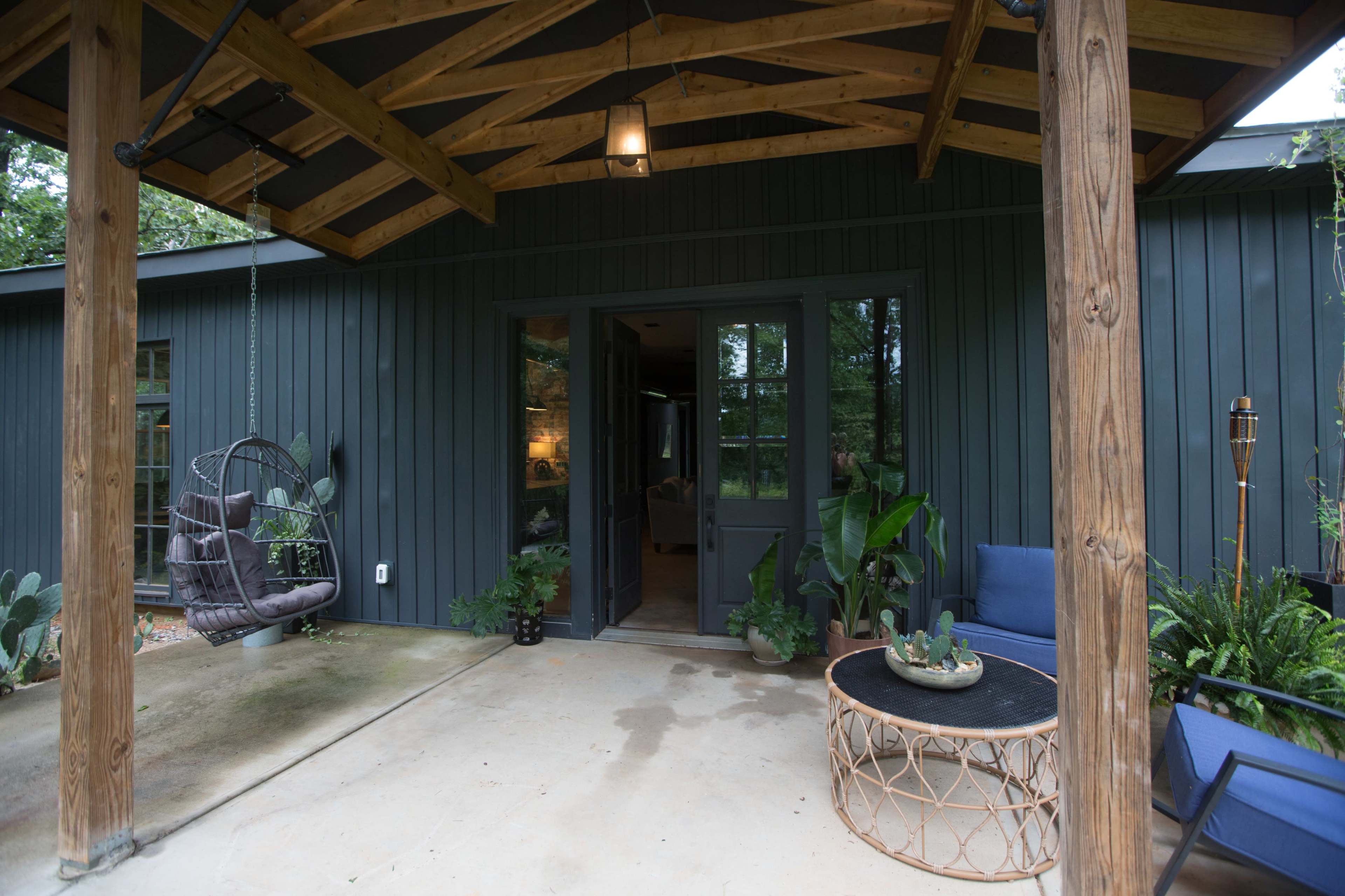 A porch with a hanging chair, a round coffee table, and two lounge chairs, set against a modern dark-clad house surrounded by greenery.