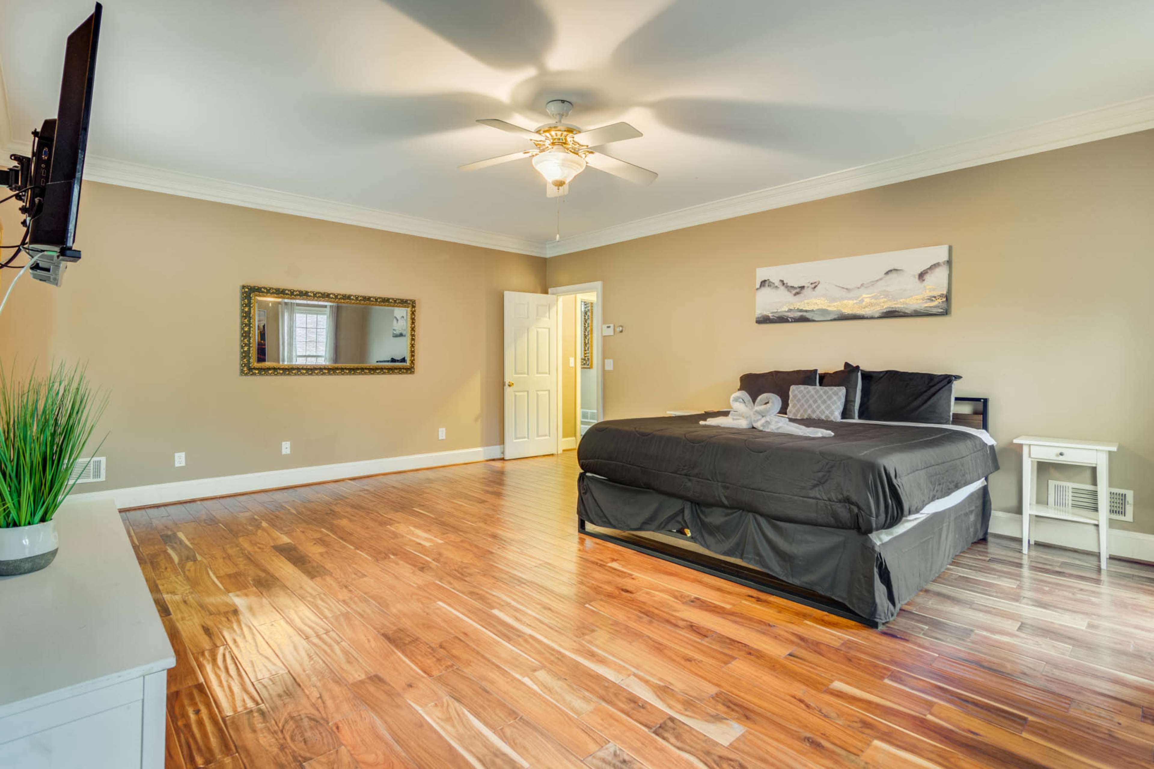 The image shows a spacious bedroom with a large bed, wooden flooring, a ceiling fan, a television mounted on the wall, and a mirror above a small table.