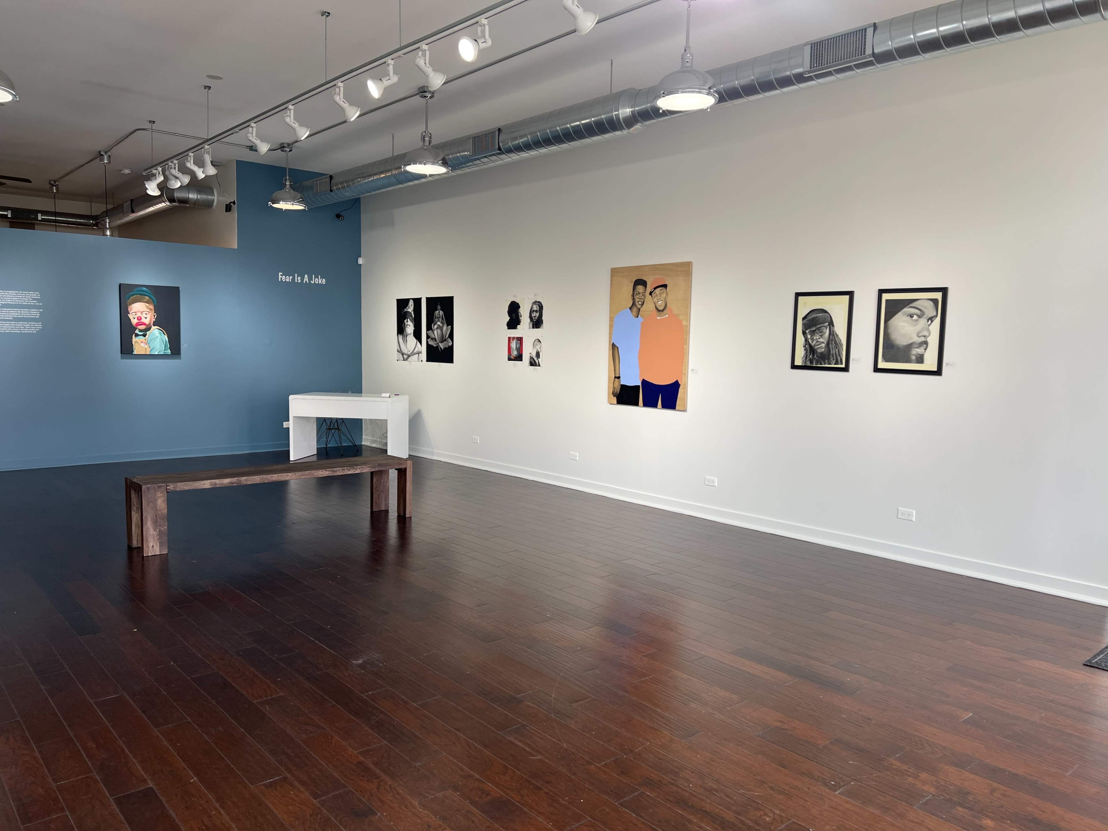 An art gallery with a wooden bench in the center, featuring a blue wall with text and several framed artworks displayed on the white walls.