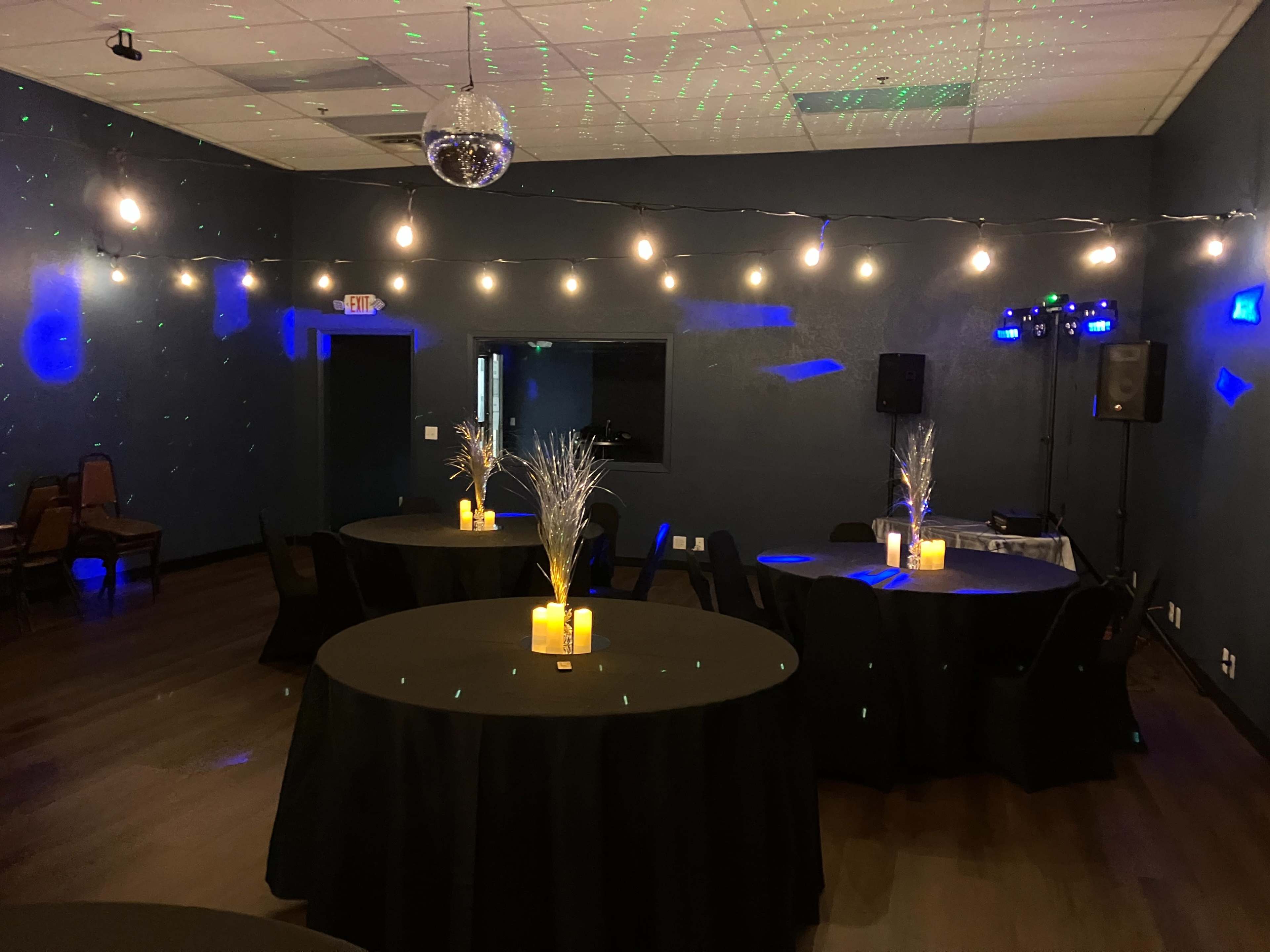 The image shows a dimly lit event space with black circular tables, each featuring a lit centerpiece and surrounded by chairs, while string lights and a disco ball add decorative elements to the ceiling.