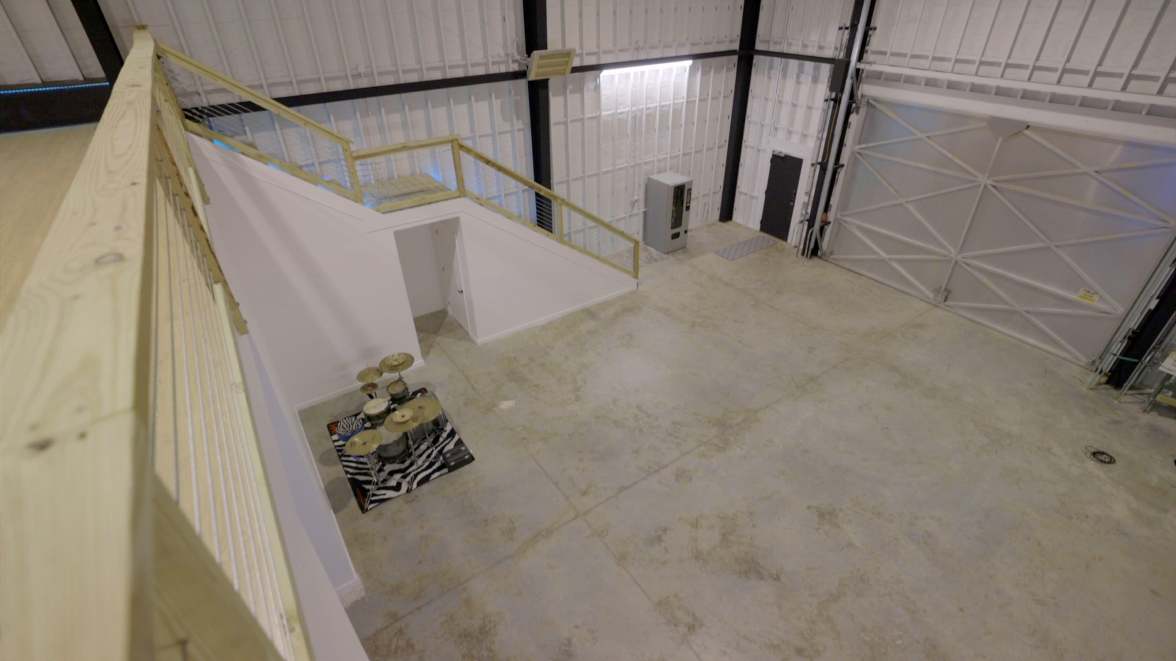 An expansive industrial space features a concrete floor, a staircase leading to a raised platform, and a few items including a drum set on a patterned mat.