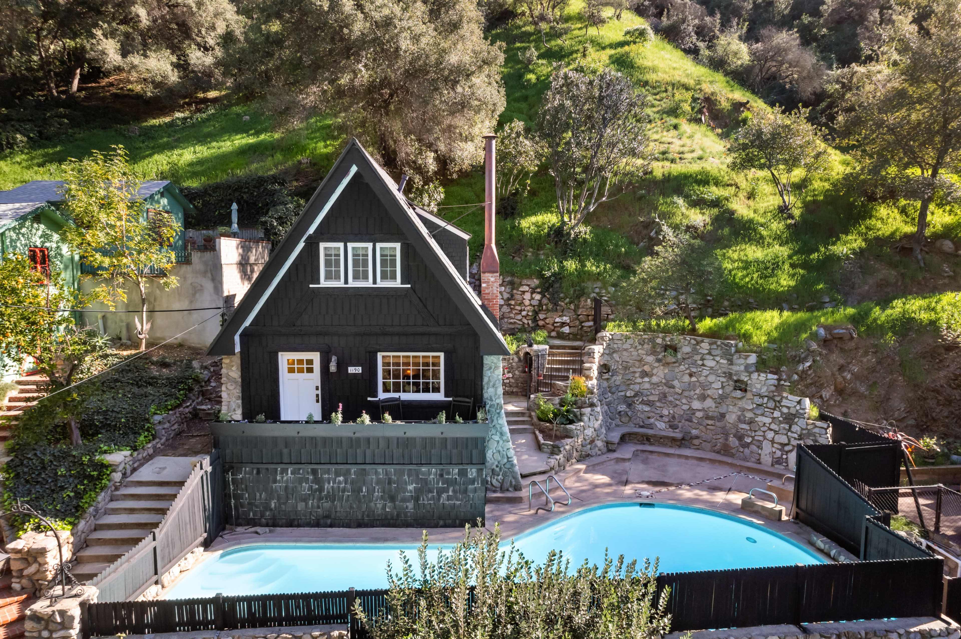 The image shows a black A-frame cabin surrounded by greenery, with a swimming pool and stone walls in the foreground.