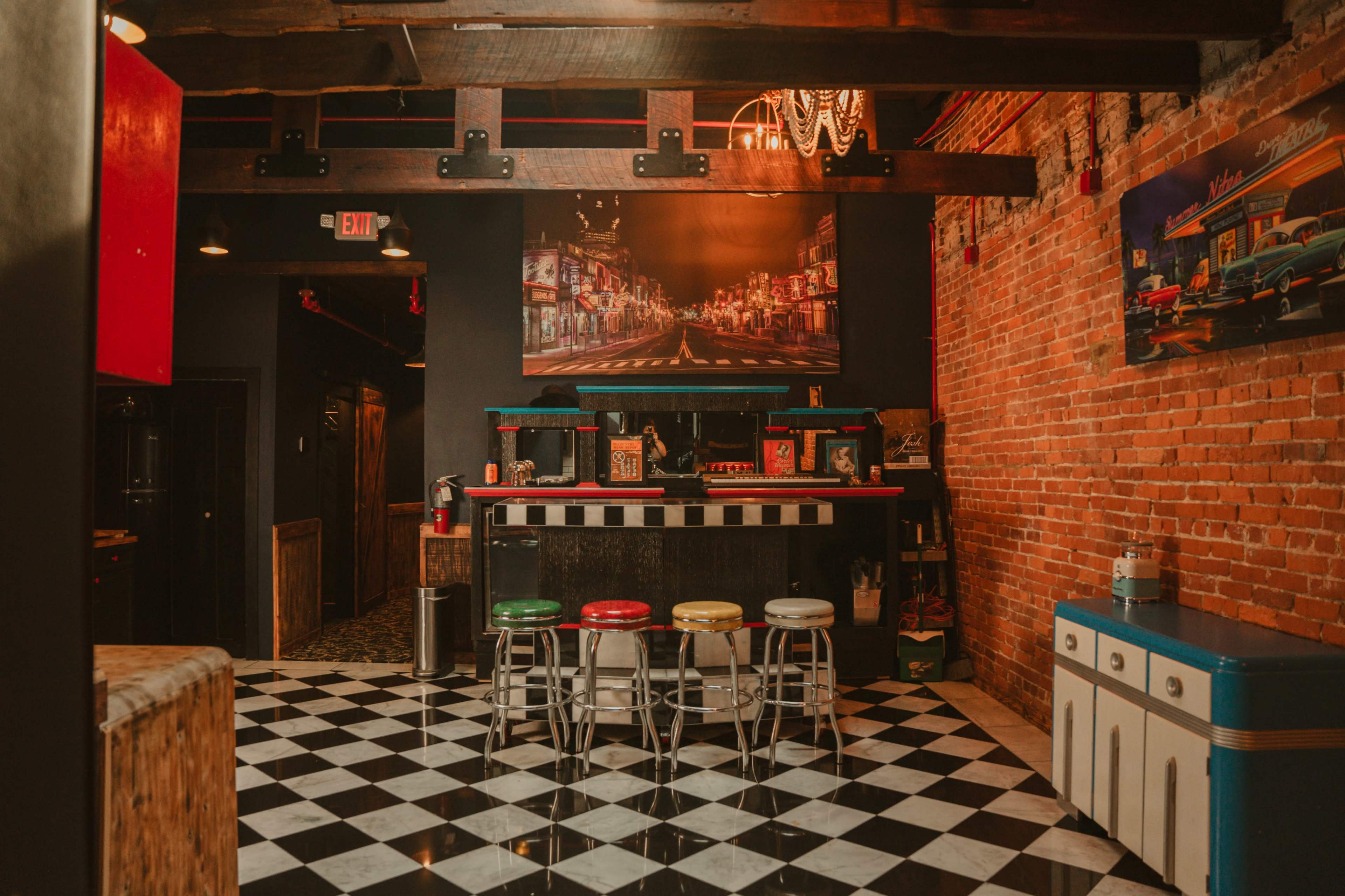 The image shows a vintage-style diner with a checkered floor, colorful stools, exposed brick walls, and a large framed photograph on the wall.
