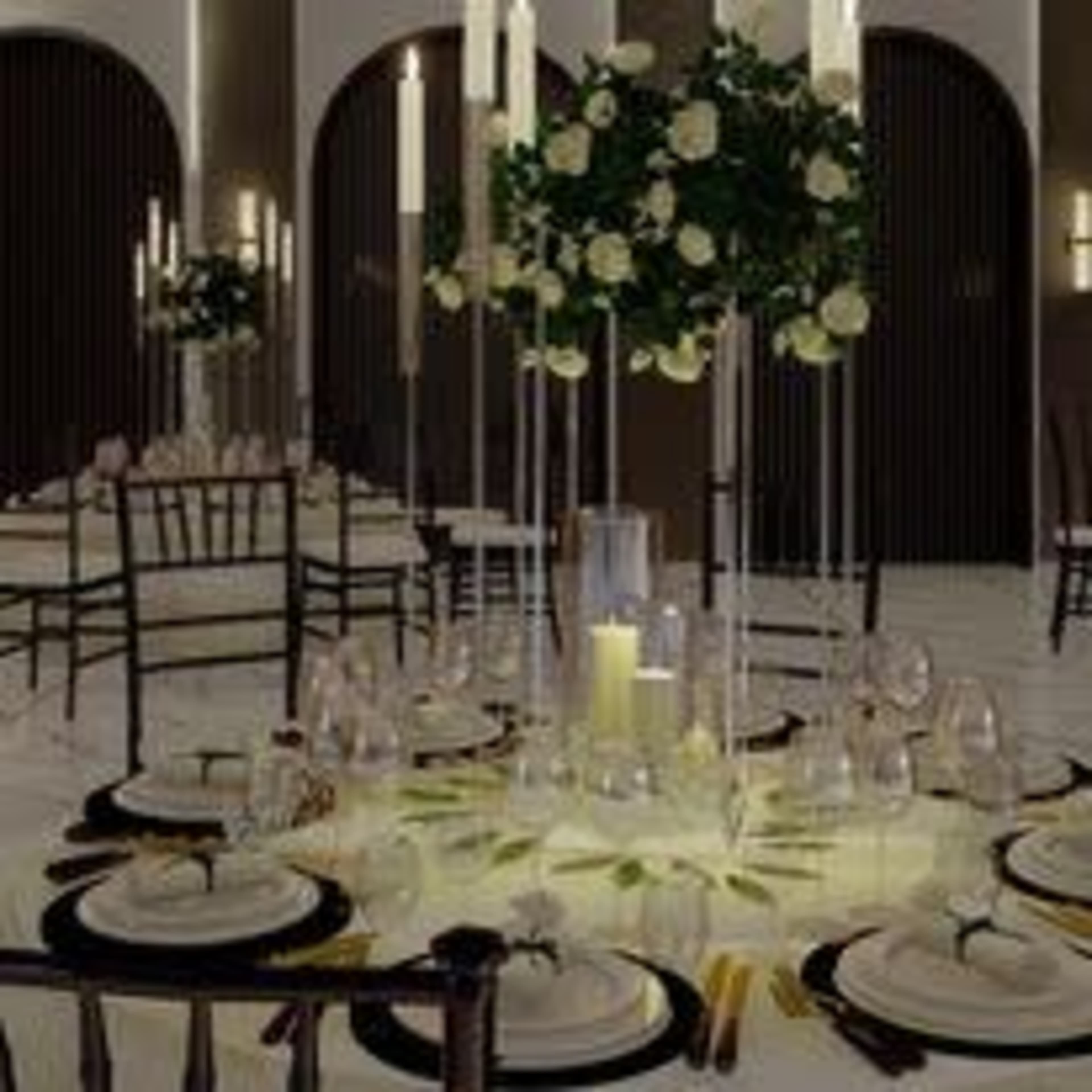 A formal dining setup with elegantly arranged tables featuring white plates, glassware, and tall candle holders, surrounded by floral centerpieces.