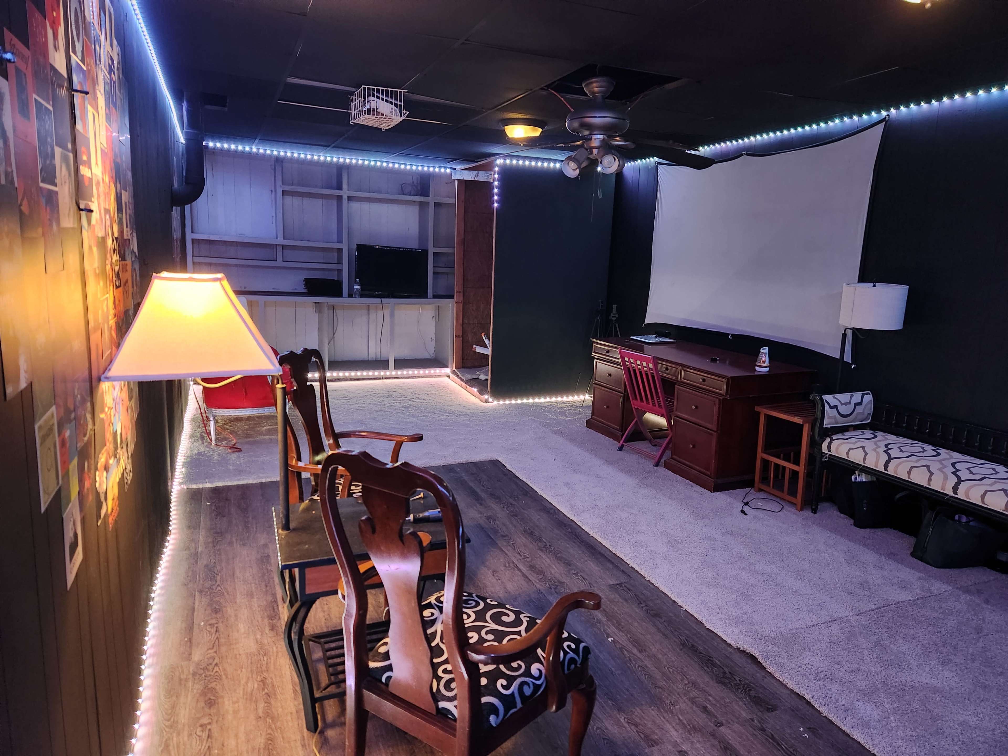 The image shows a basement room furnished with chairs, a desk, and a wall-mounted screen, illuminated by LED lights along the perimeter.