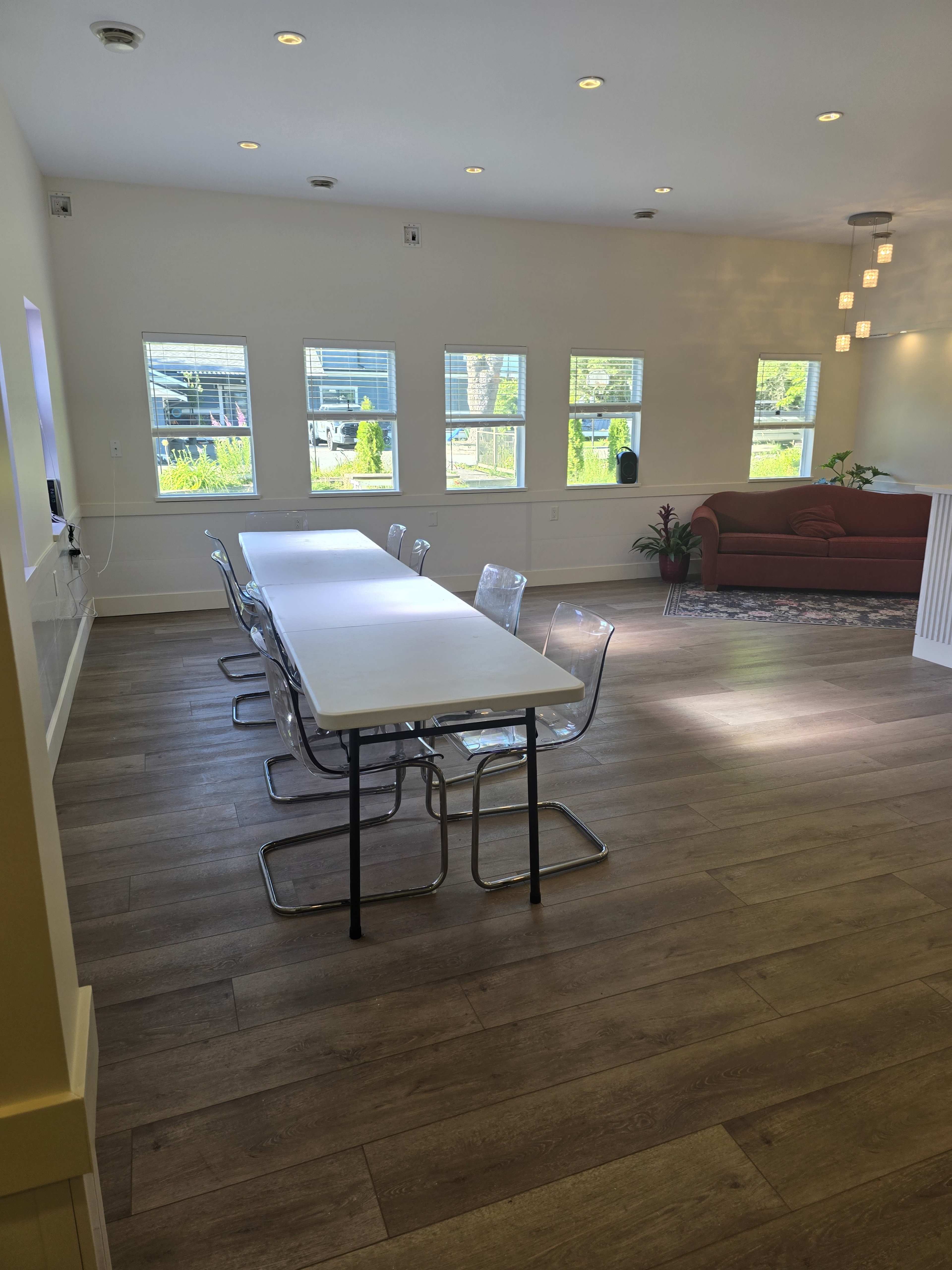 The image shows a bright, modern room with a long white table and clear chairs, illuminated by natural light from large windows, and a red couch in the background.