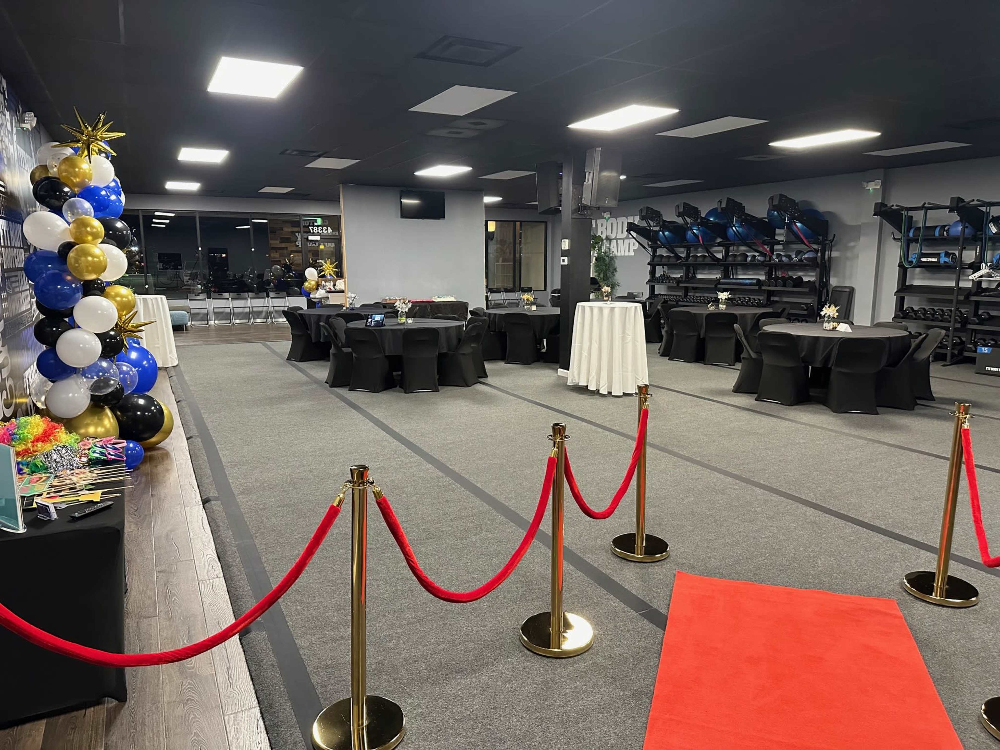 The image shows a dimly lit event space featuring decorated tables, a red carpet with stanchions, and a backdrop of balloons.