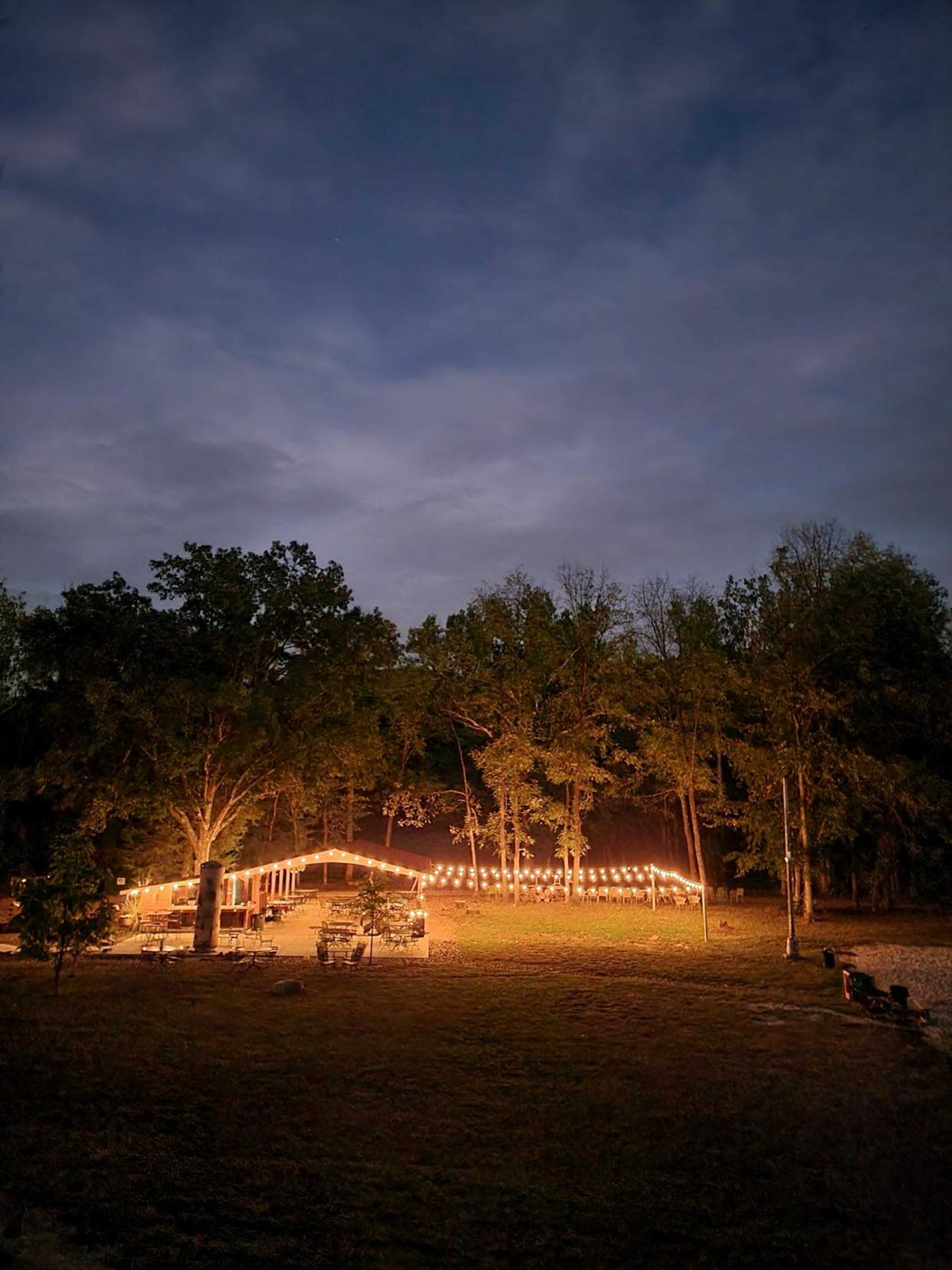 A warmly lit structure is nestled among trees under a cloudy evening sky.