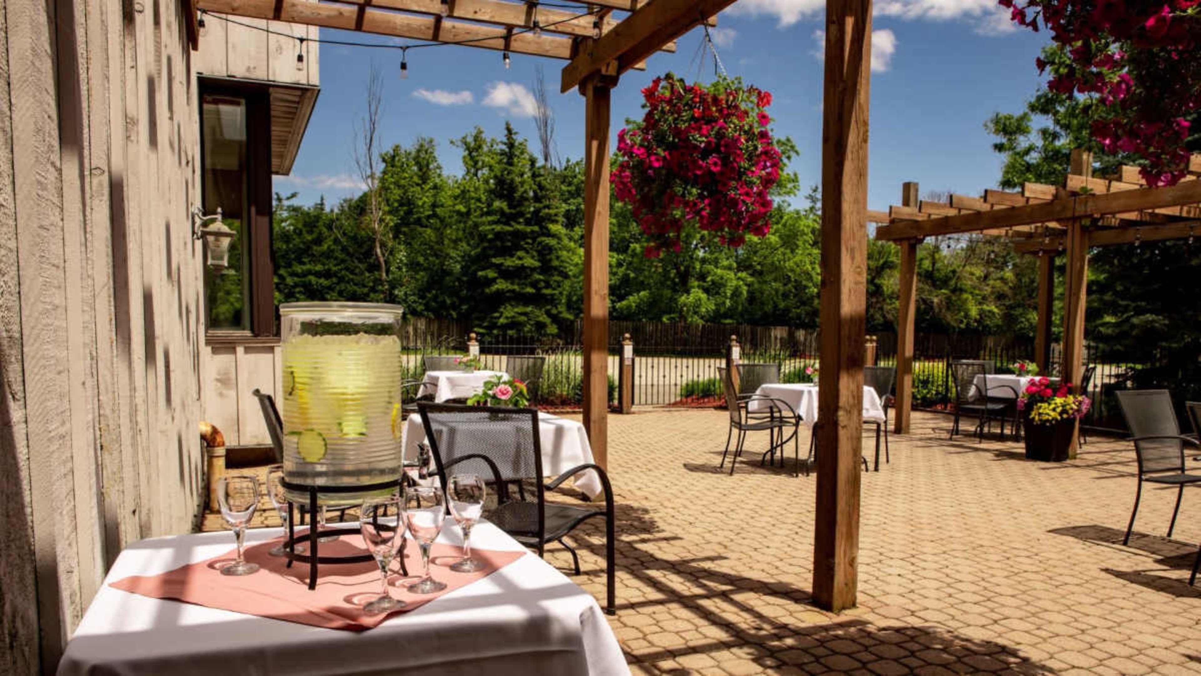 The image shows a patio area of a restaurant with tables set for dining, surrounded by greenery and hanging flower baskets.