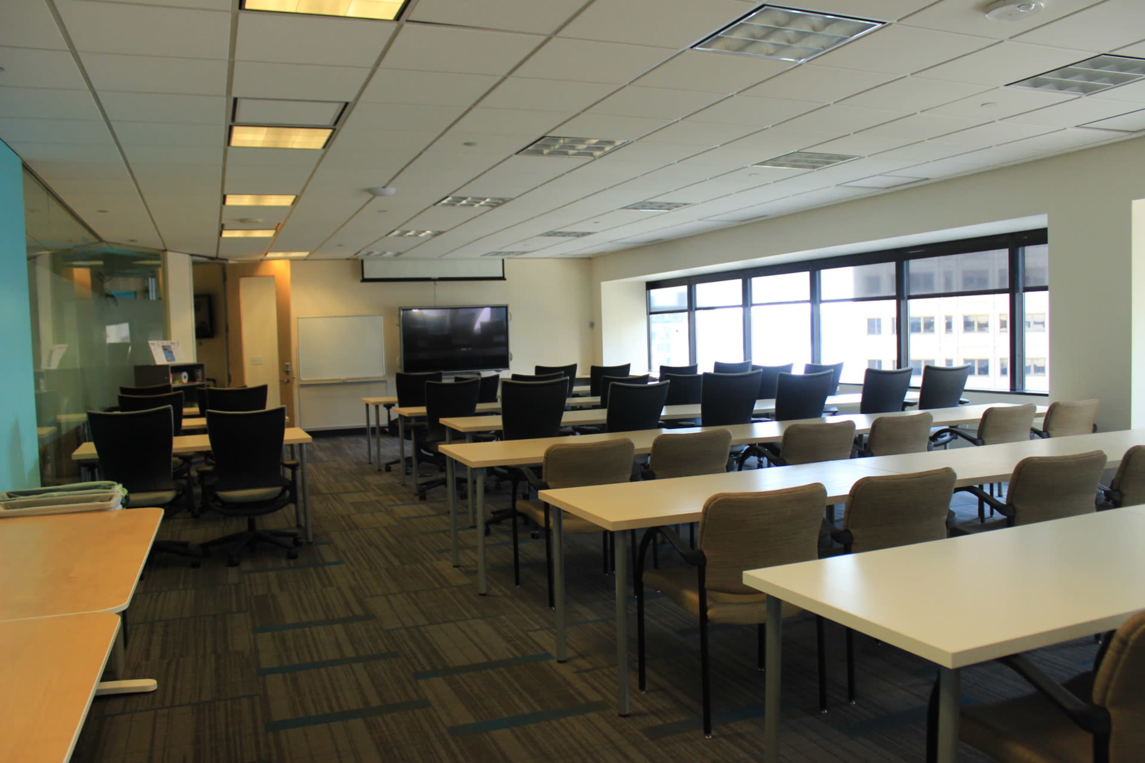 A modern conference room with rows of tables and chairs, large windows, and a screen for presentations.