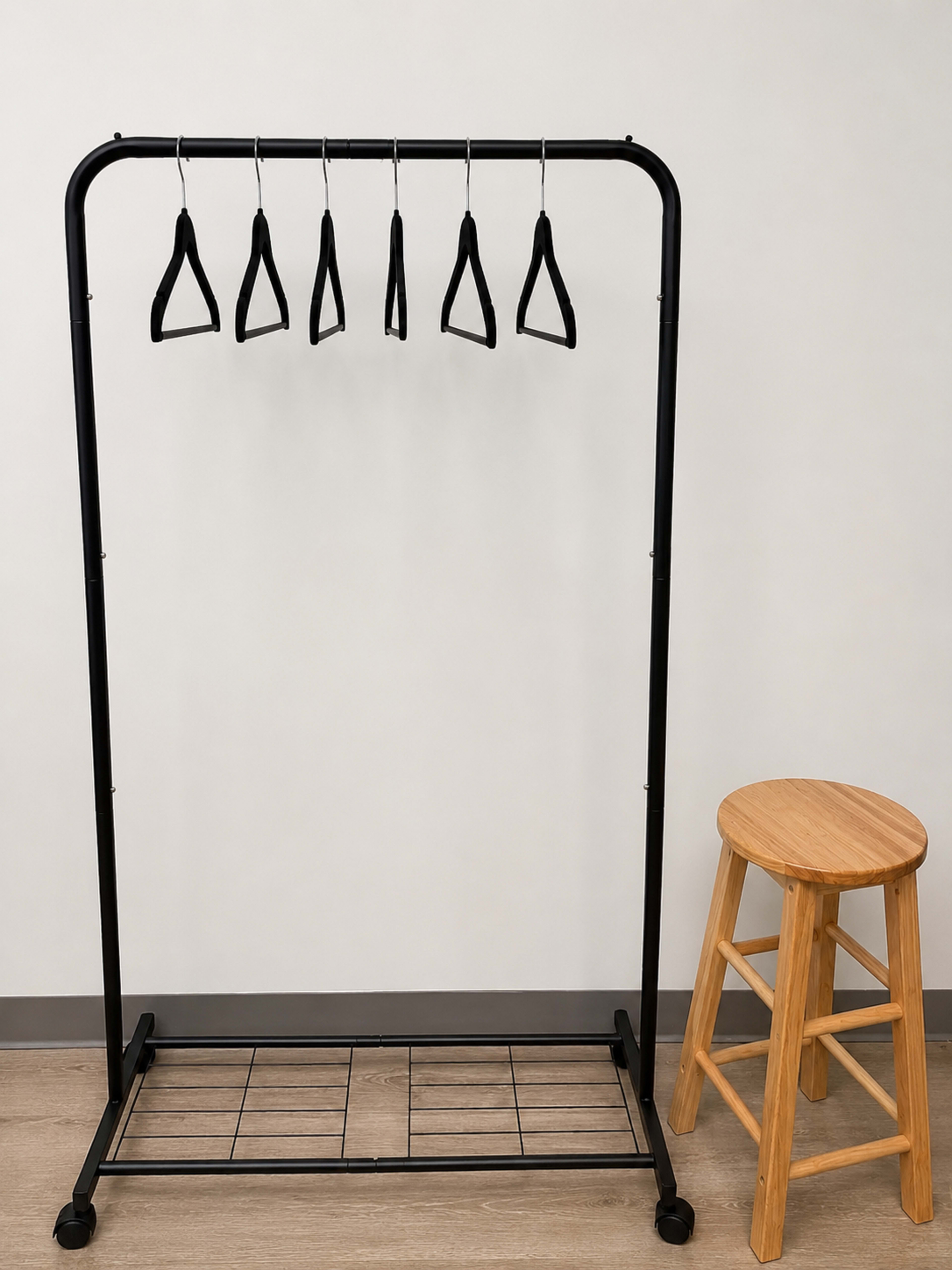 A black clothing rack with five hangers is positioned next to a wooden stool against a light-colored wall.