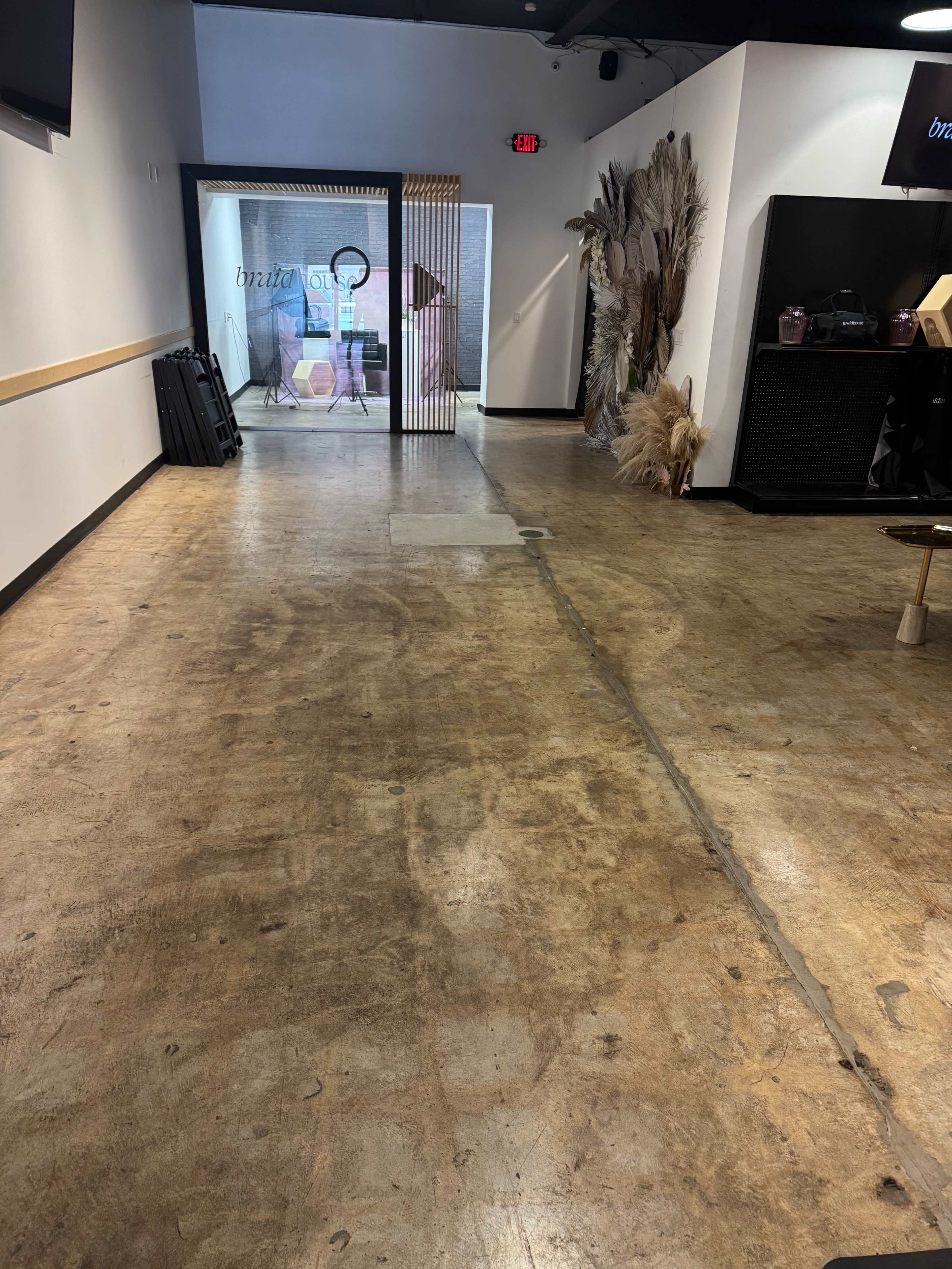 The image shows a hallway with a concrete floor, leading to a doorway framed by decorative elements, with some plants and furniture visible in the background.