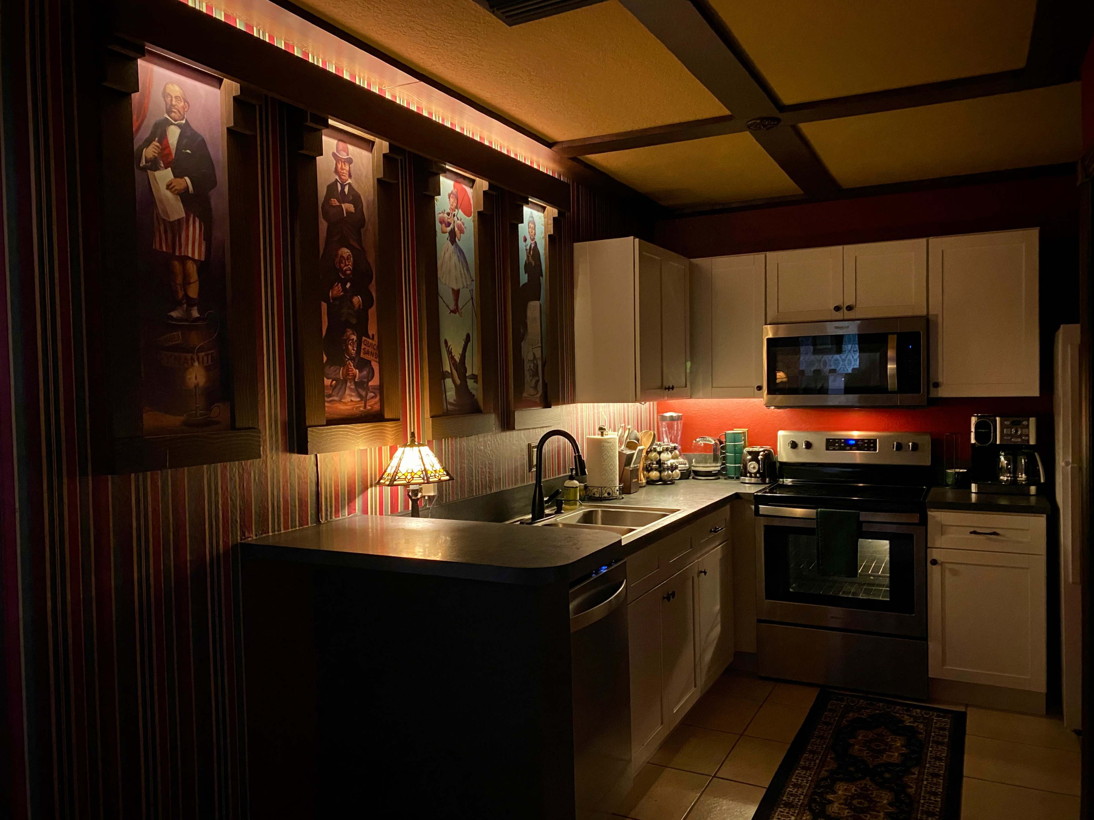 The image shows a dimly lit kitchen featuring white cabinets, stainless steel appliances, and artwork on the walls.