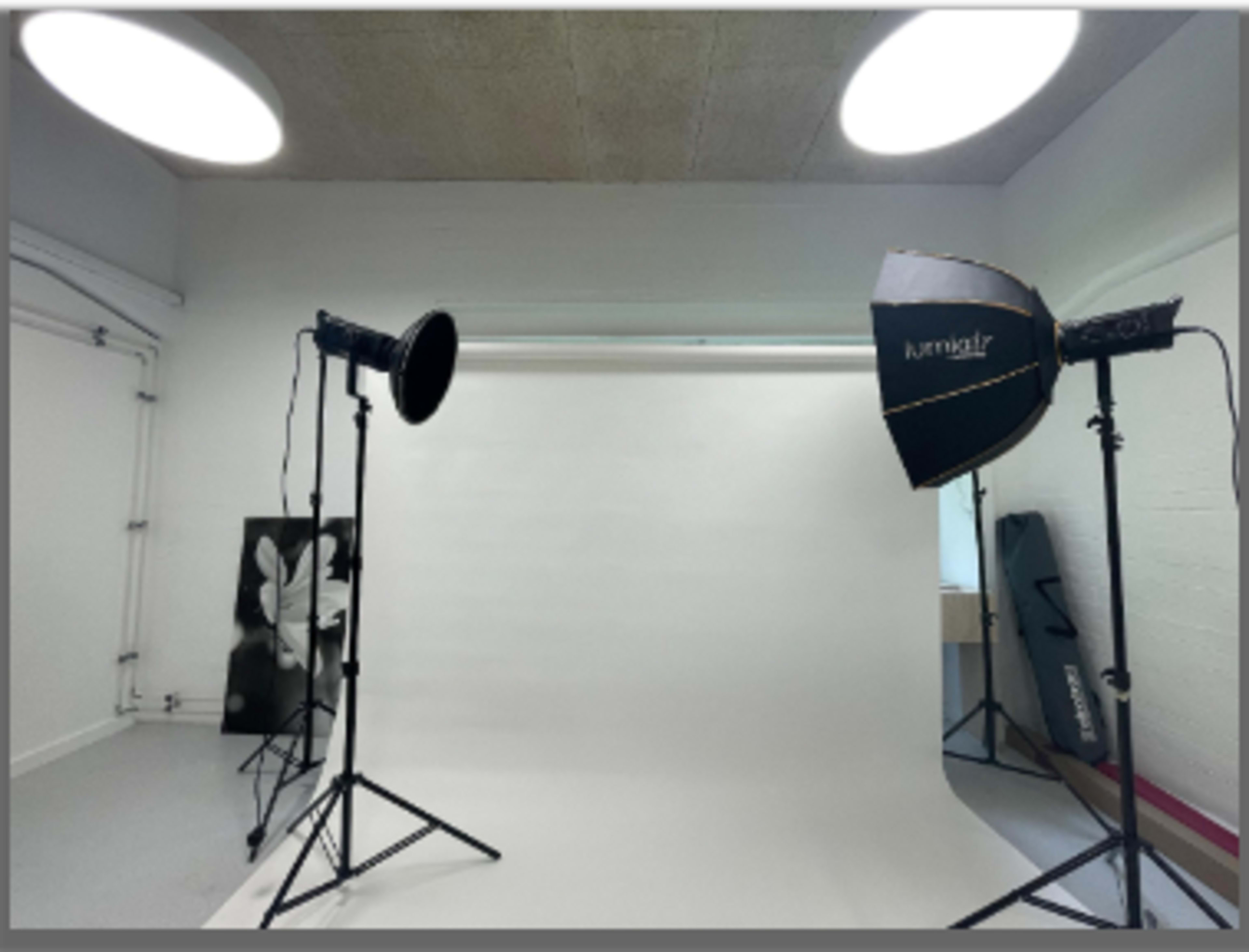 A photography studio with a white backdrop, two softbox lights on stands, and equipment in the corner.