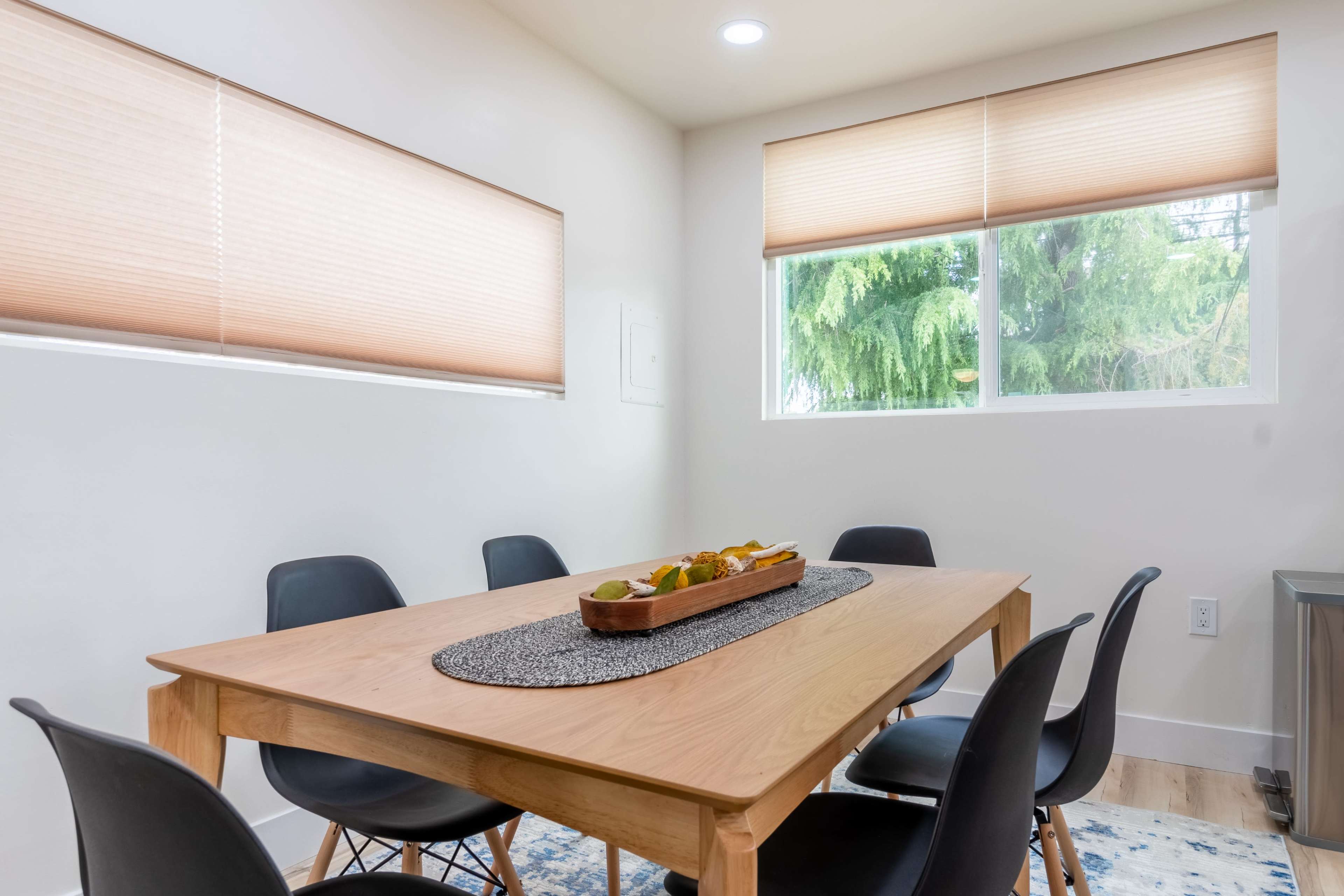 A wooden dining table surrounded by black chairs is set in a bright room with two windows featuring beige blinds.