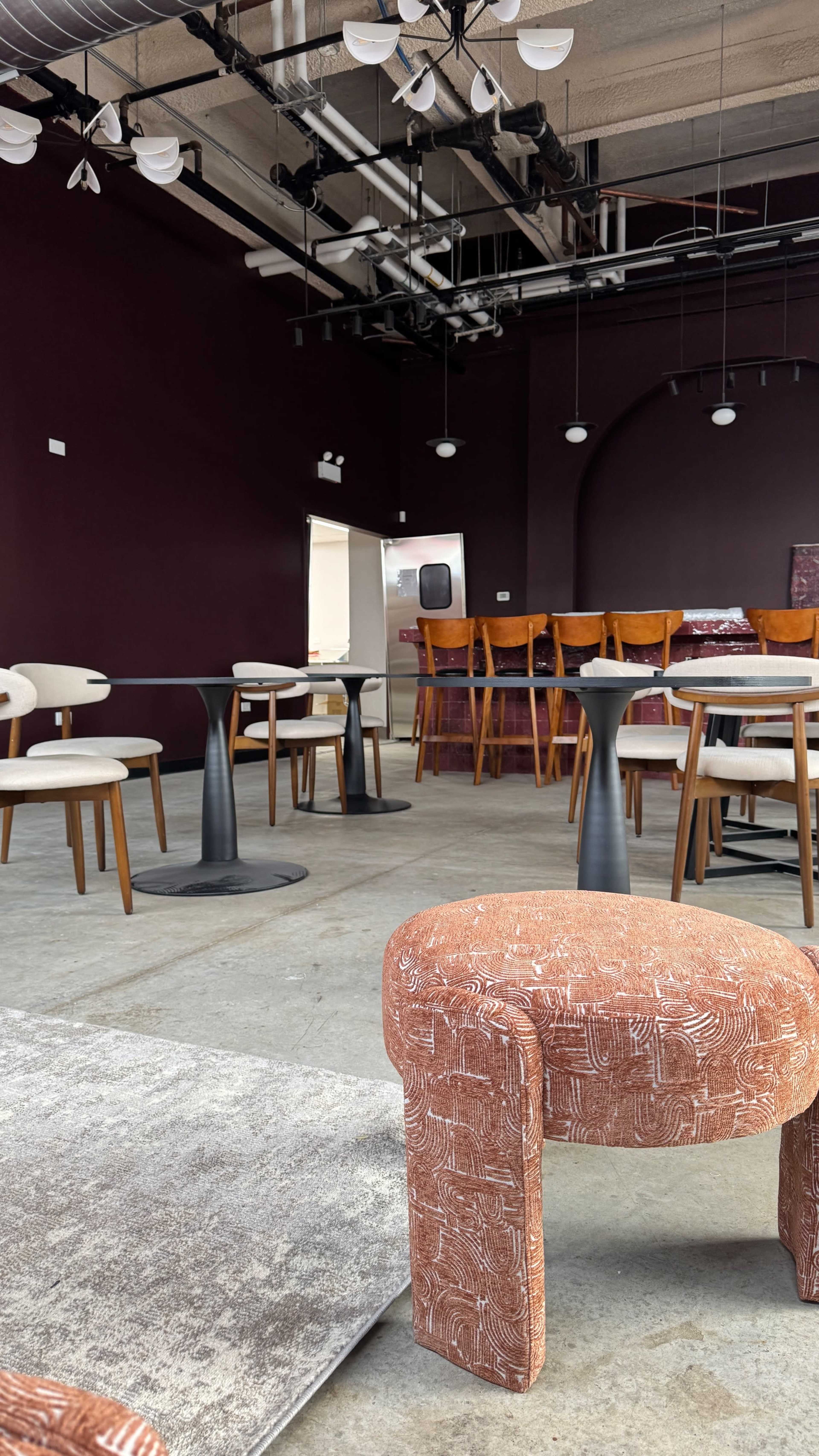The image shows a modern café interior with a mix of small tables and chairs, a large bar area in the background, and a maroon-colored wall.