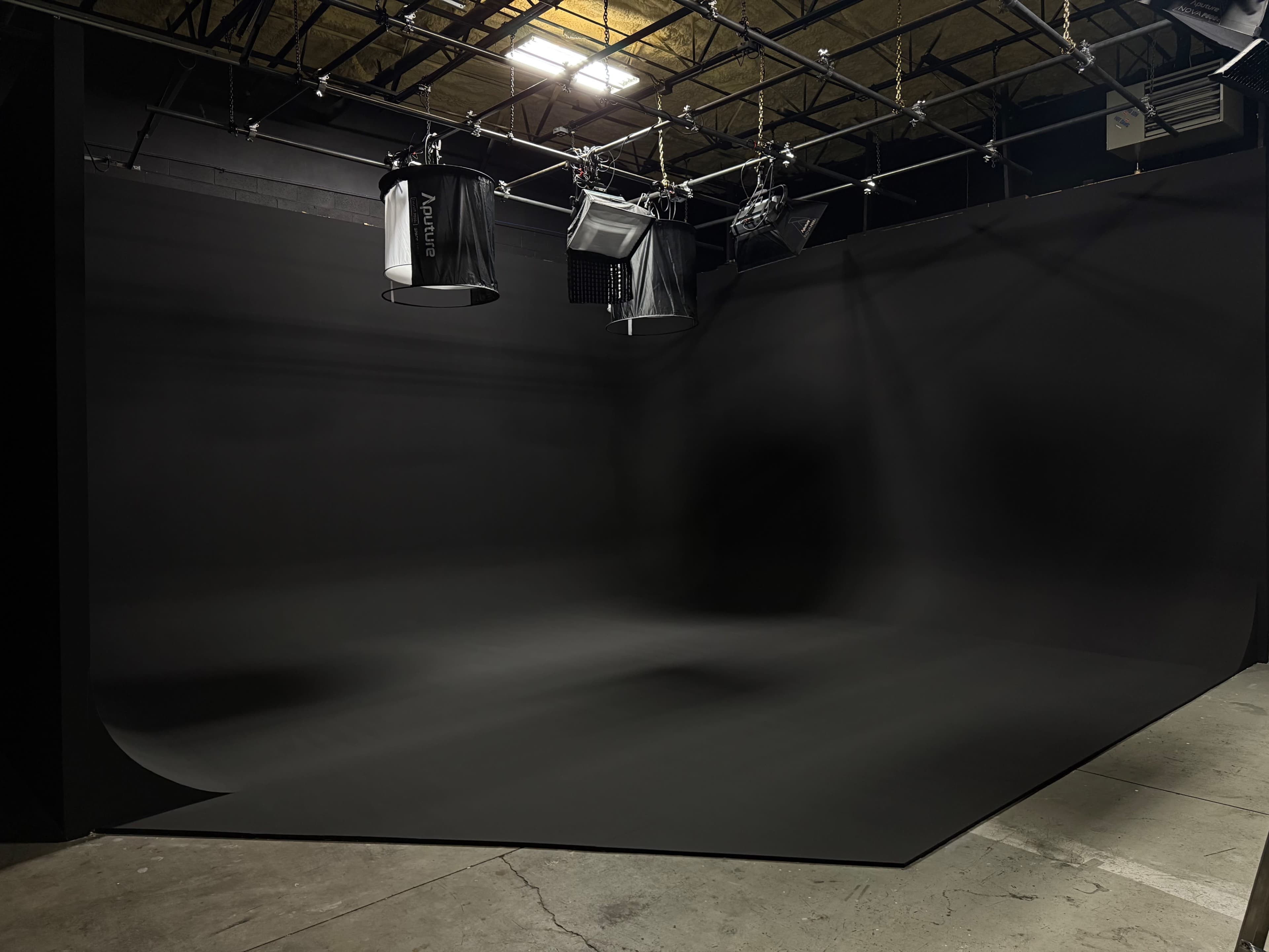 The image depicts a photography studio featuring a large black backdrop and various lighting equipment suspended from the ceiling.