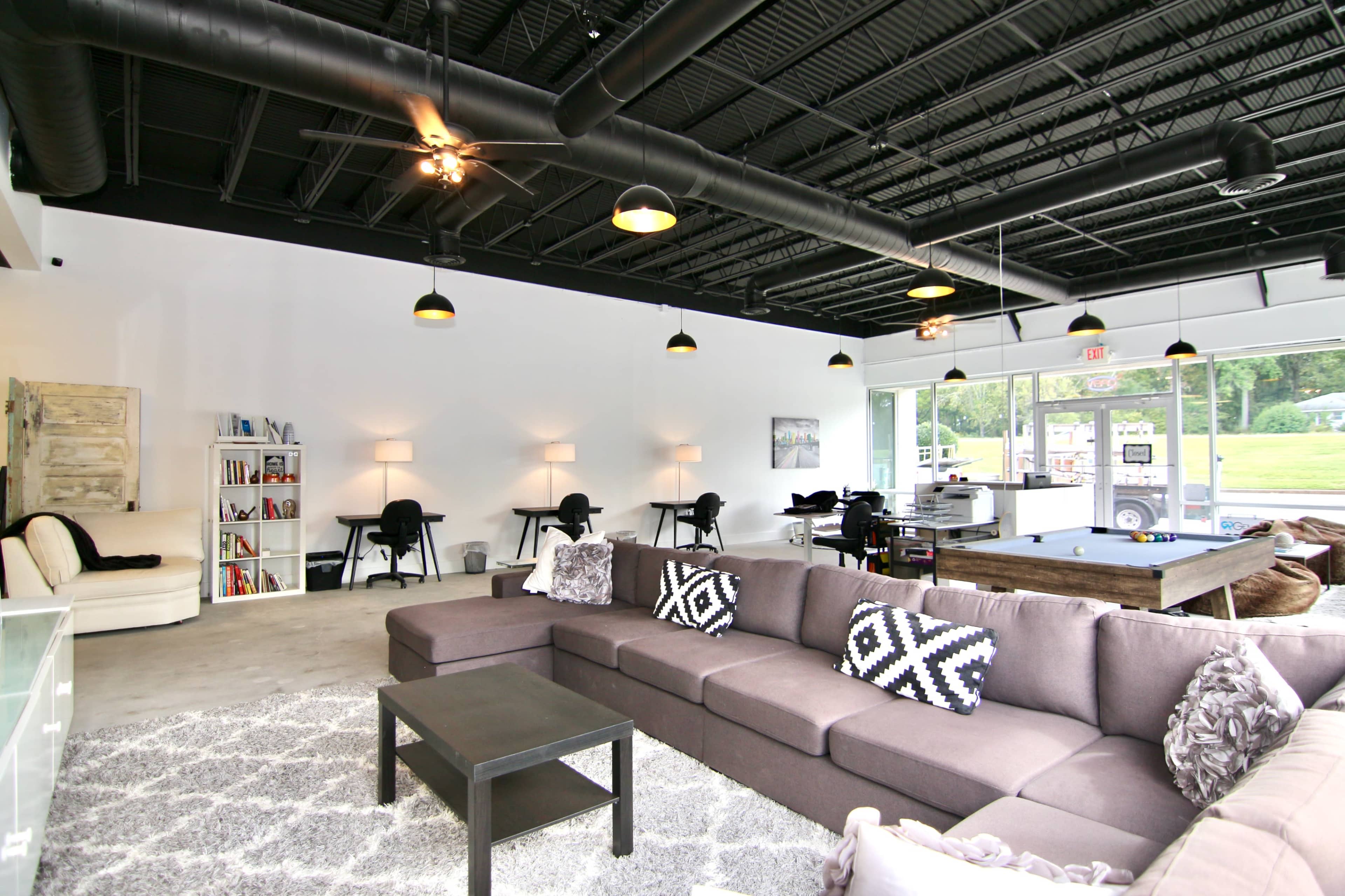 A modern lounge area with a large gray sectional sofa, a coffee table, and several workspaces, all surrounded by large windows and a pool table in the background.