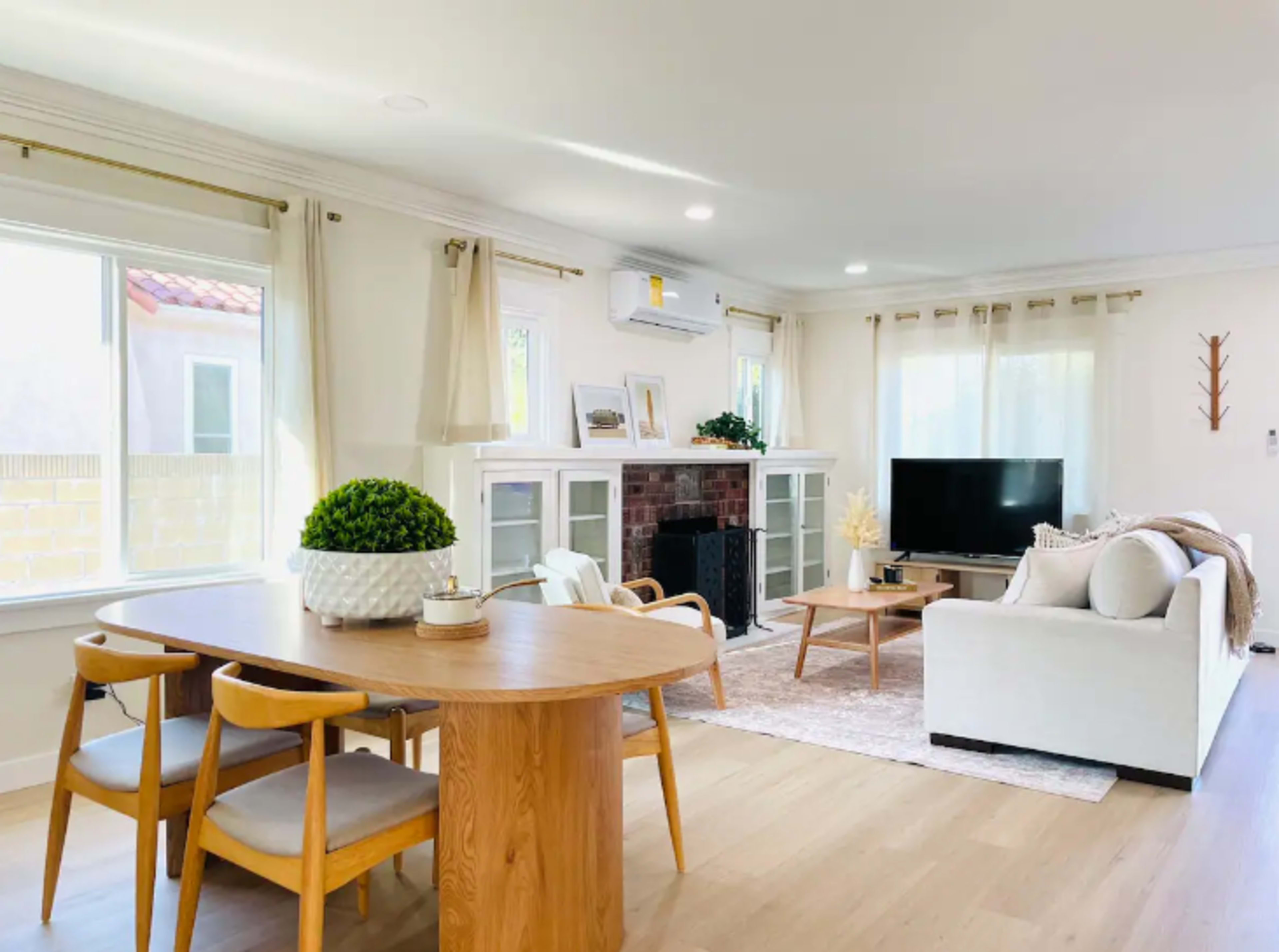 A modern living room with a white sofa, a wooden dining table, a television on a stand, and decorative plants.