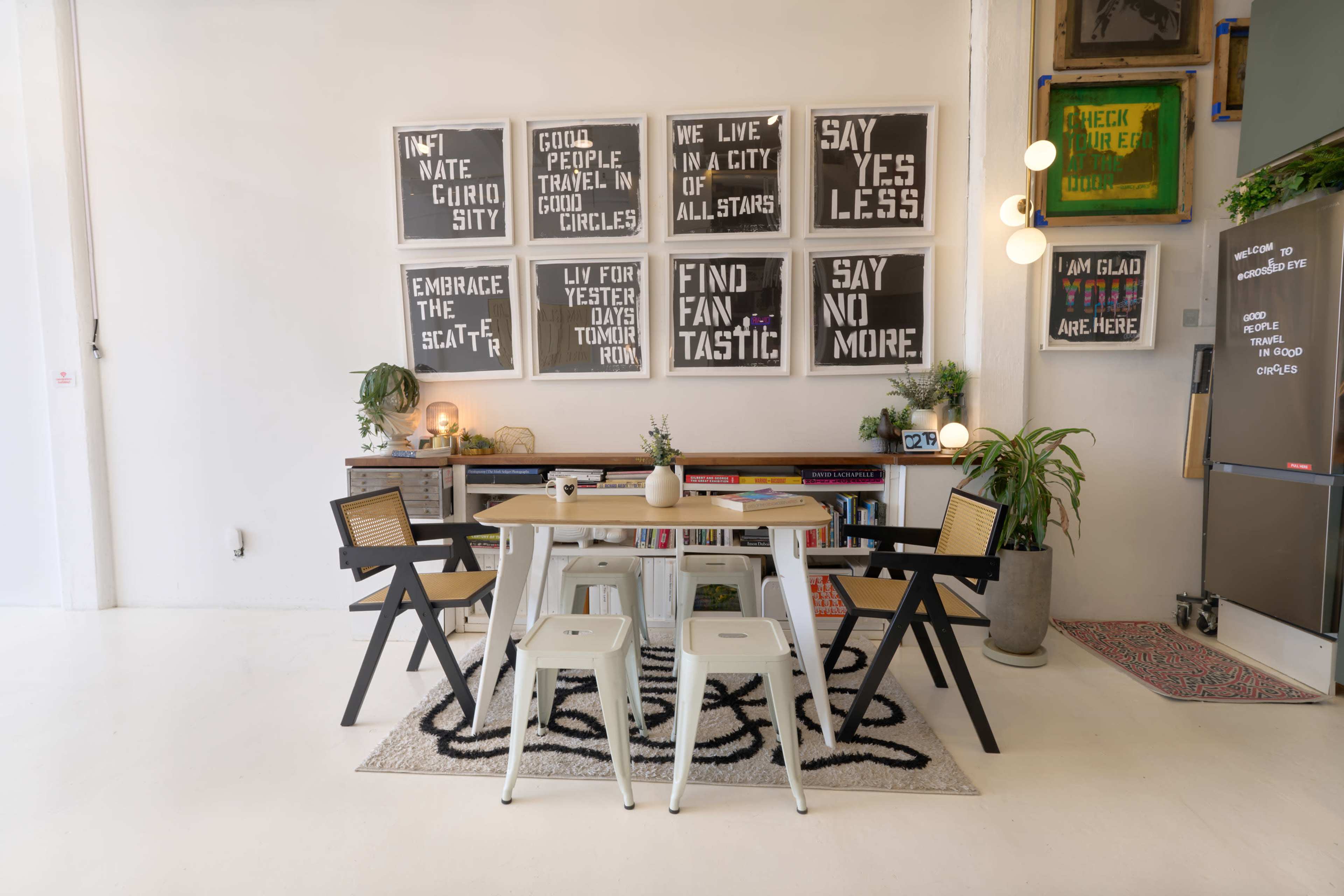A modern workspace features a table surrounded by chairs, with inspirational wall art displayed above and plants adding to the decor.
