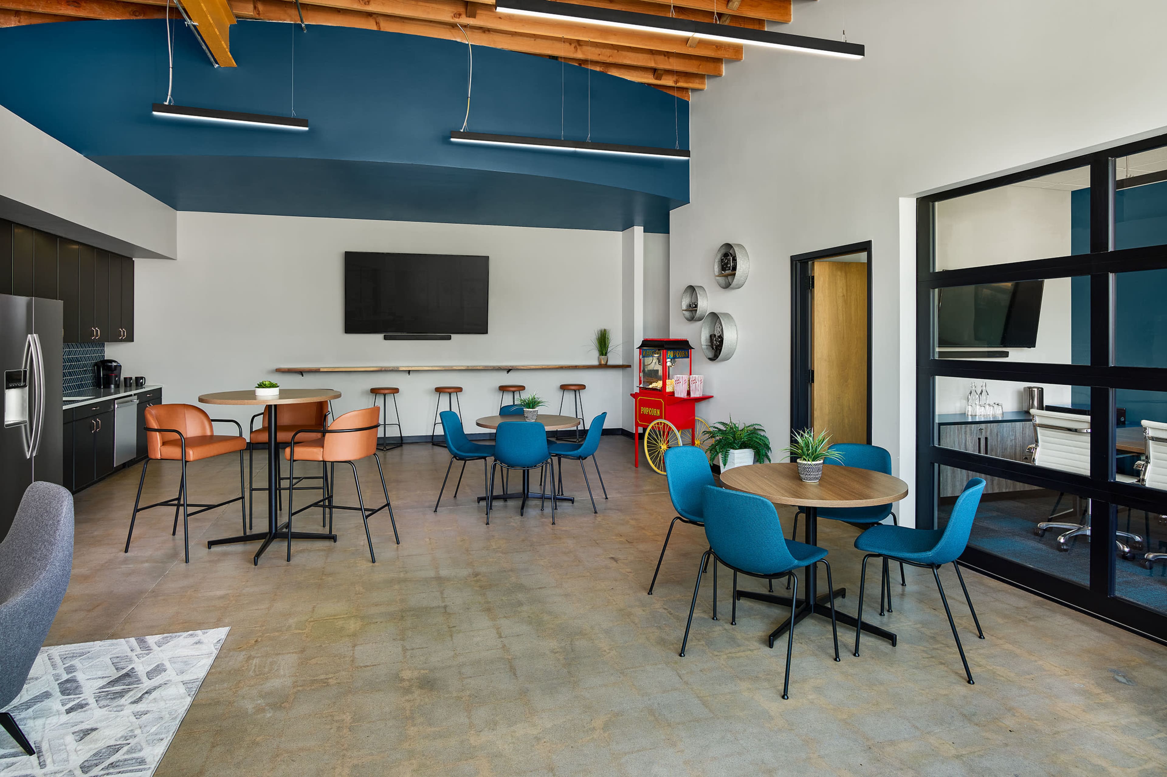 A modern office break room with a kitchenette, seating area, and a large screen mounted on the wall.