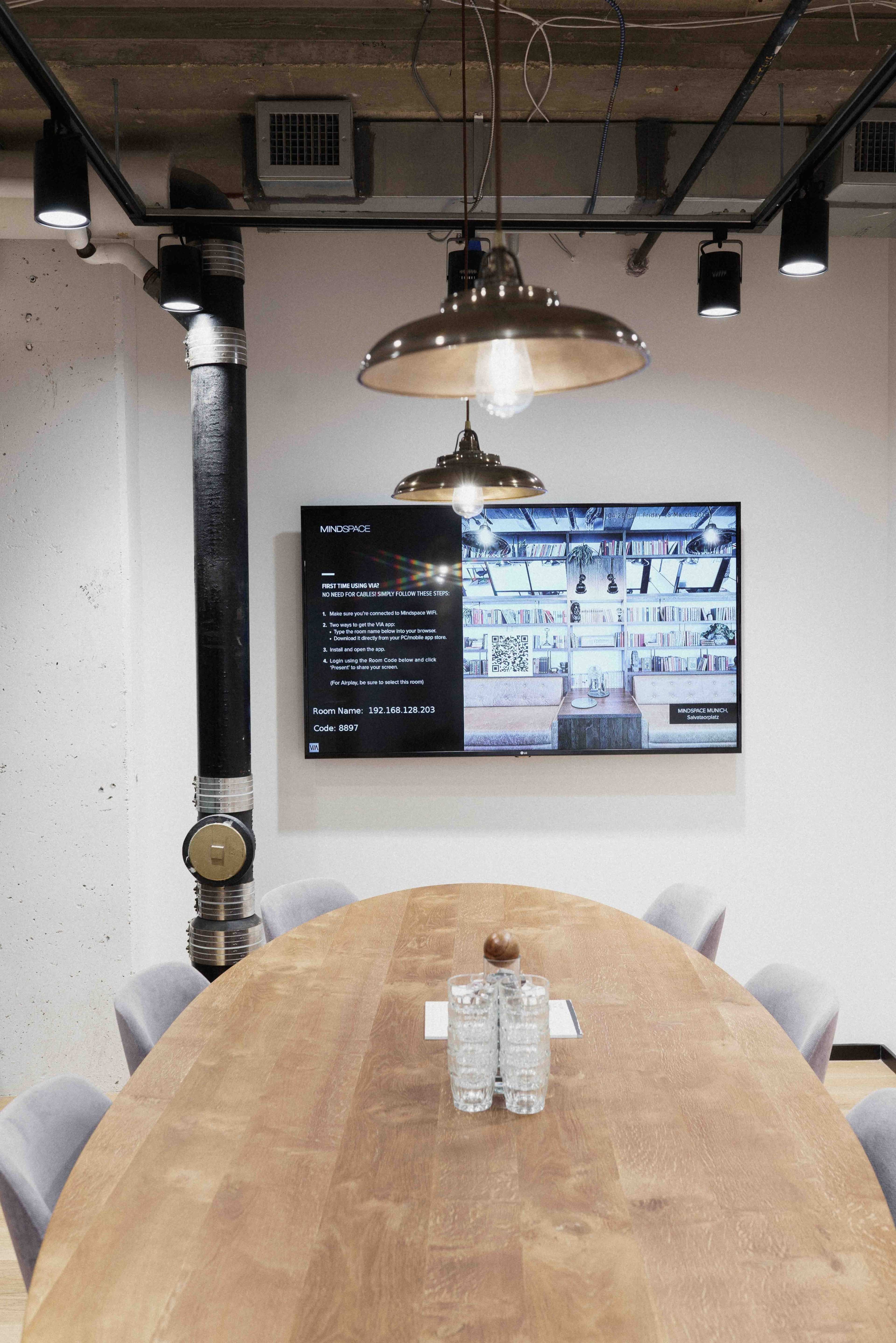 A modern conference room with a wooden table, gray chairs, and a large screen displaying information on a wall.
