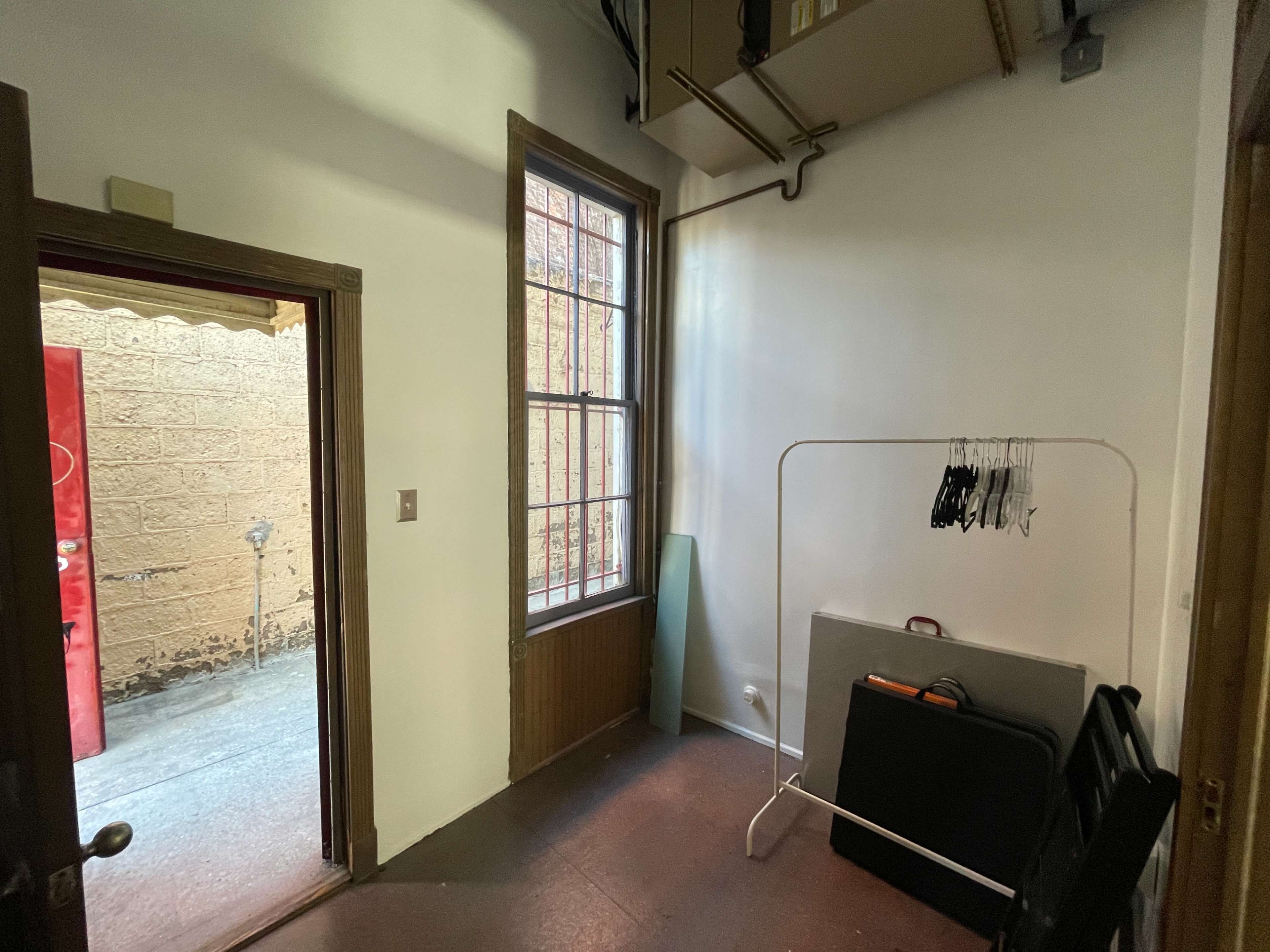 The image shows a small, sparsely furnished room with a doorway, a large window, a clothing rack with hangers, and some stacked items against the wall.