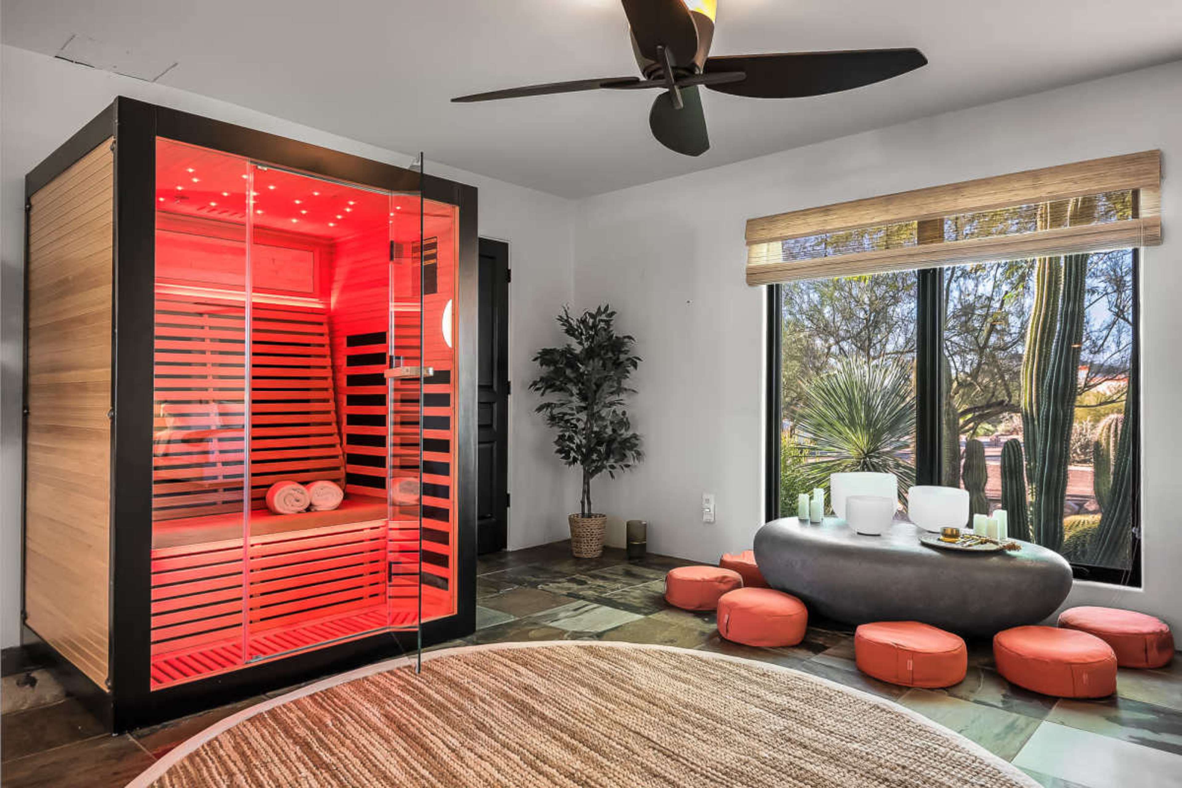 The image shows a modern sauna with red lighting beside a cozy seating area featuring low, round cushions and a stone table.