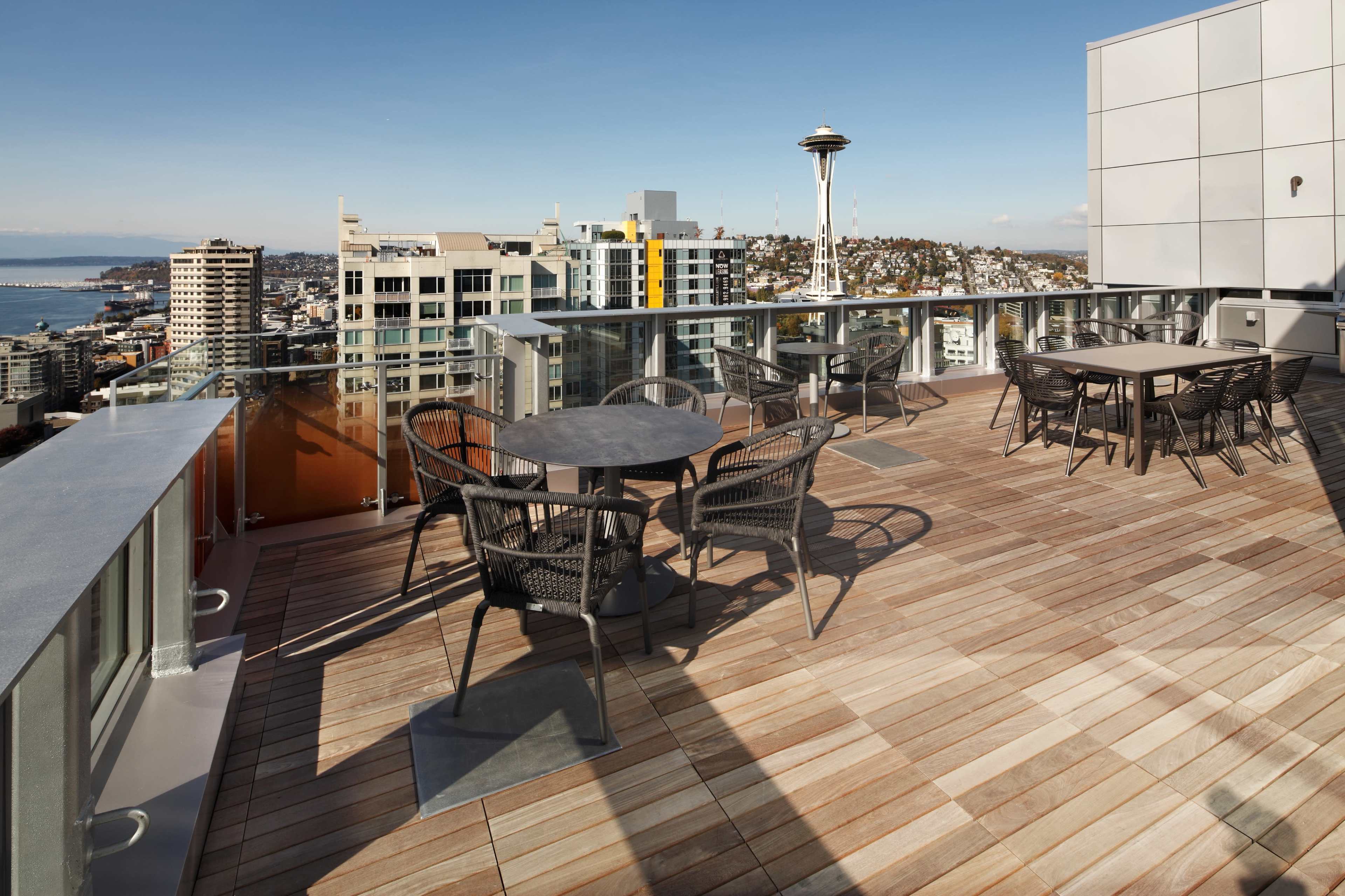 A rooftop deck with multiple tables and chairs overlooks a city skyline featuring the Space Needle in the distance.