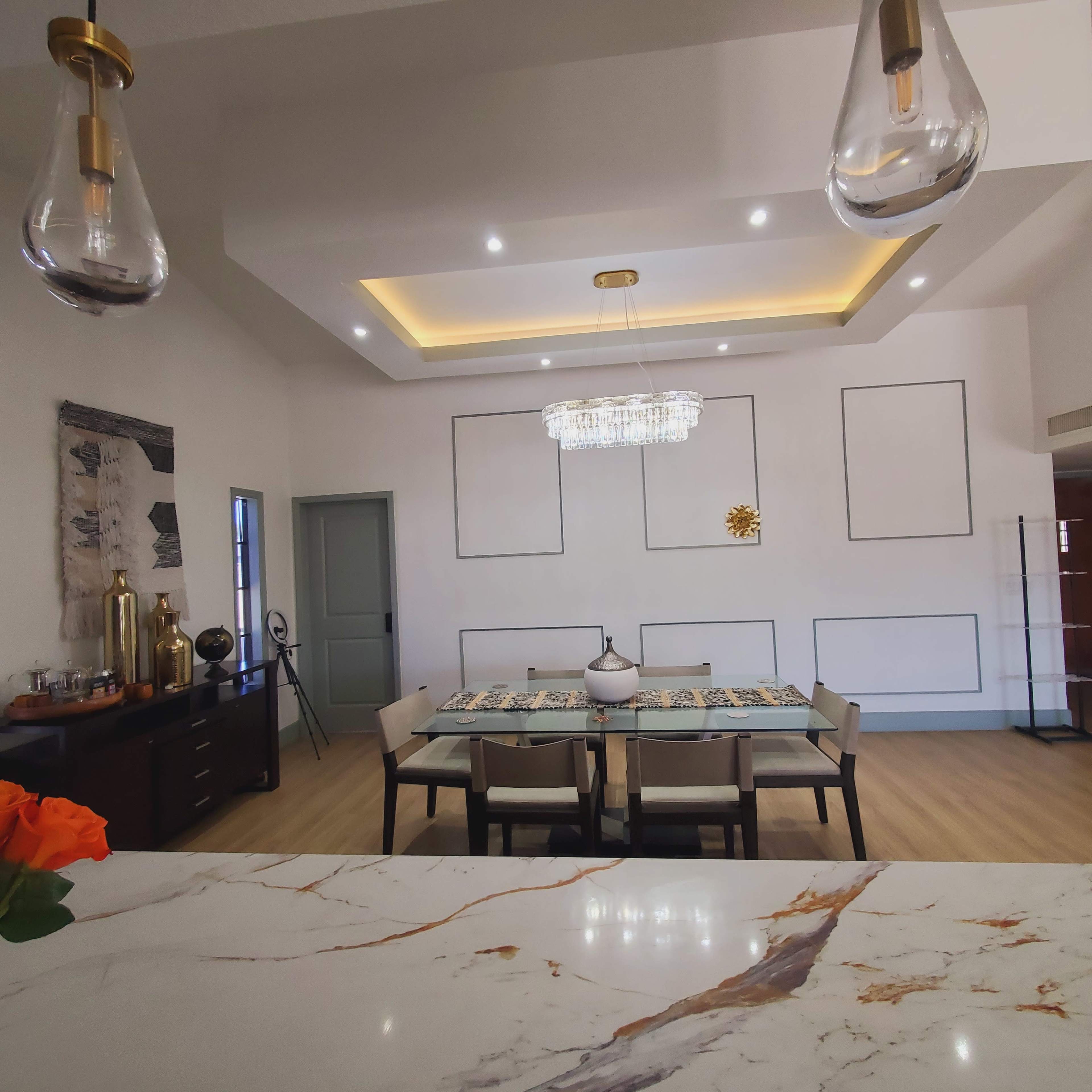 A modern dining room features a rectangular table surrounded by chairs, a large chandelier above, and decorative wall panels.